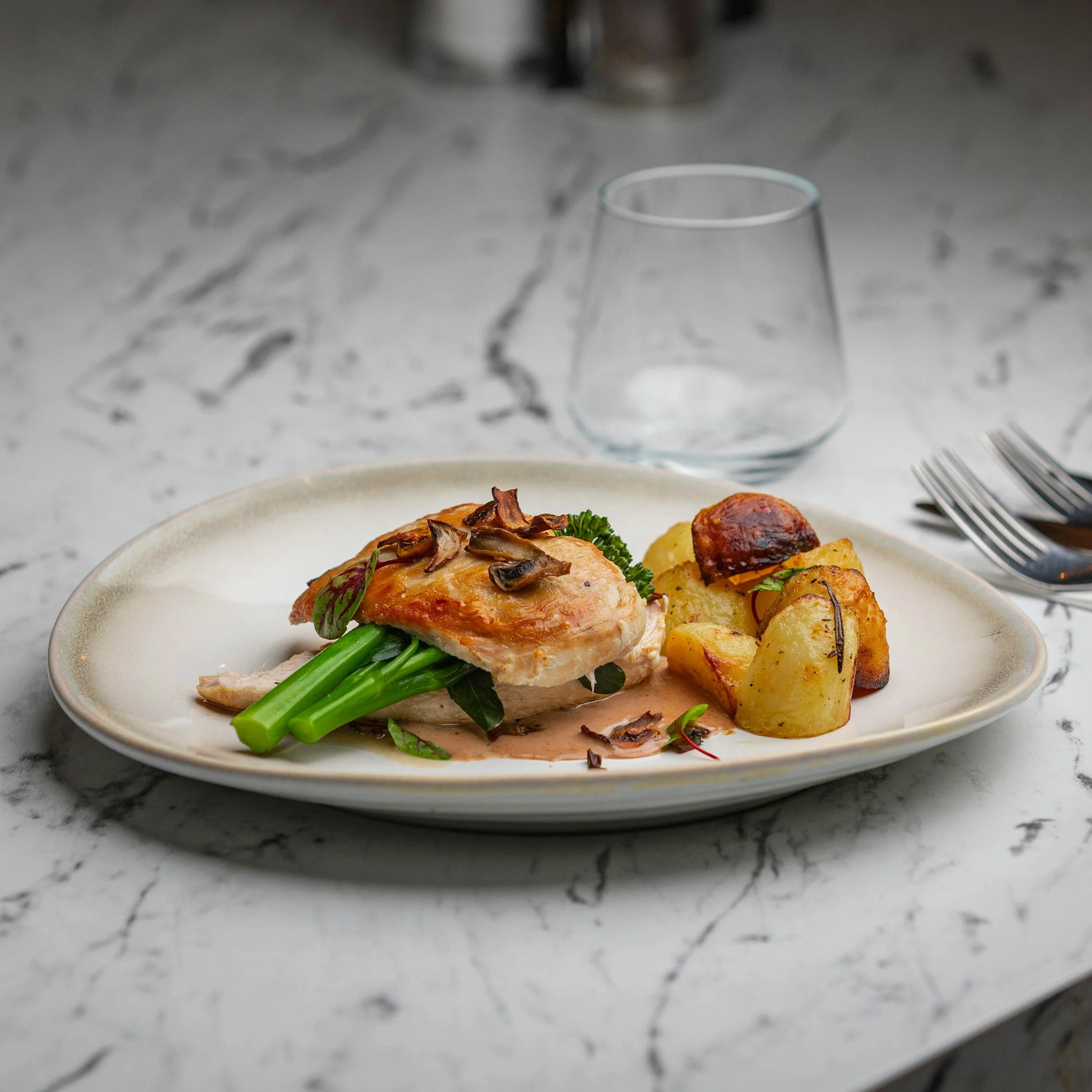 A plate with cooked chicken breast topped with mushrooms, served with green vegetables and roasted potatoes on a white plate, marble countertop, empty glass, and silverware.