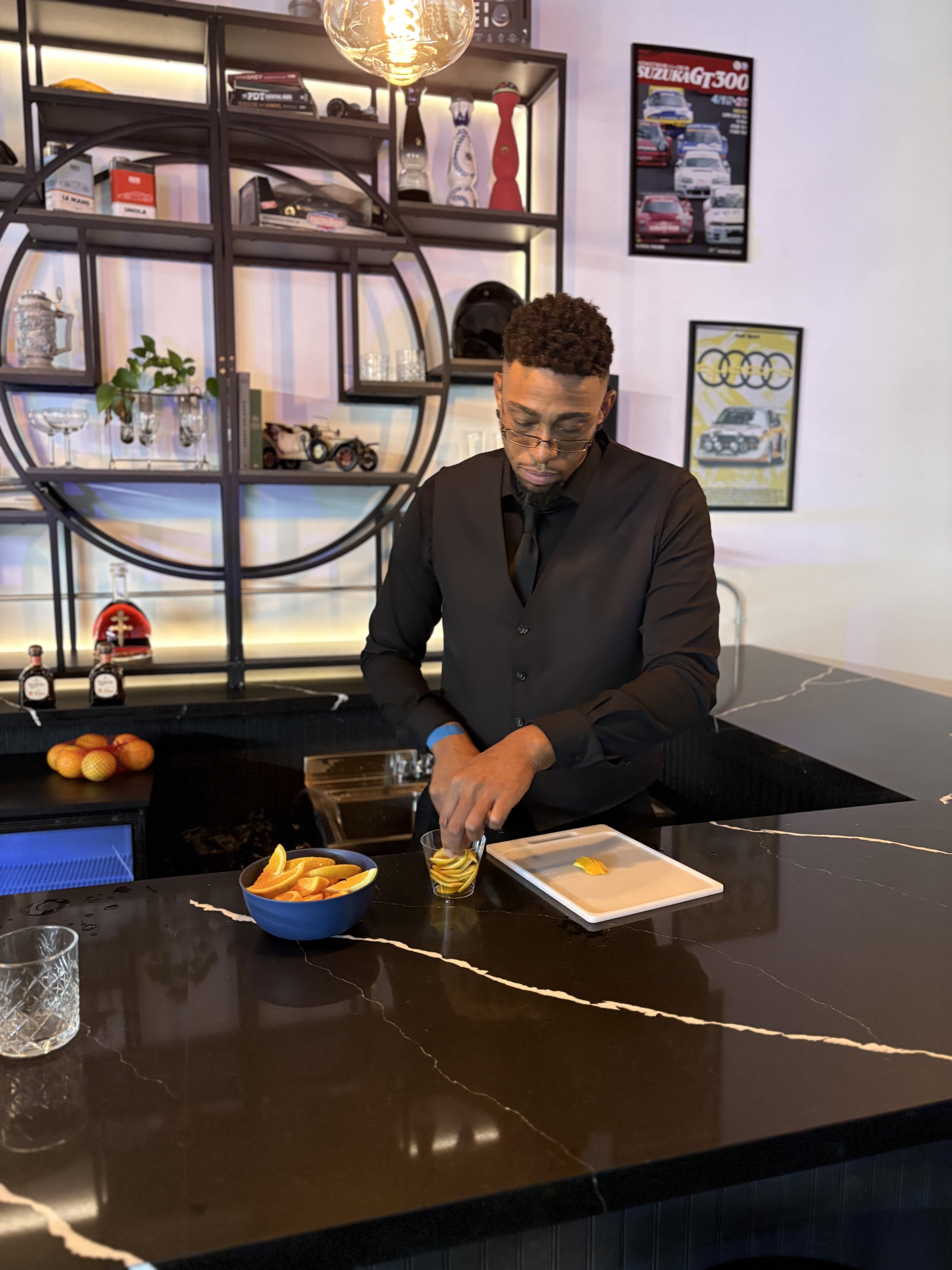 A man in black attire preparing orange slices at a black marble bar counter.