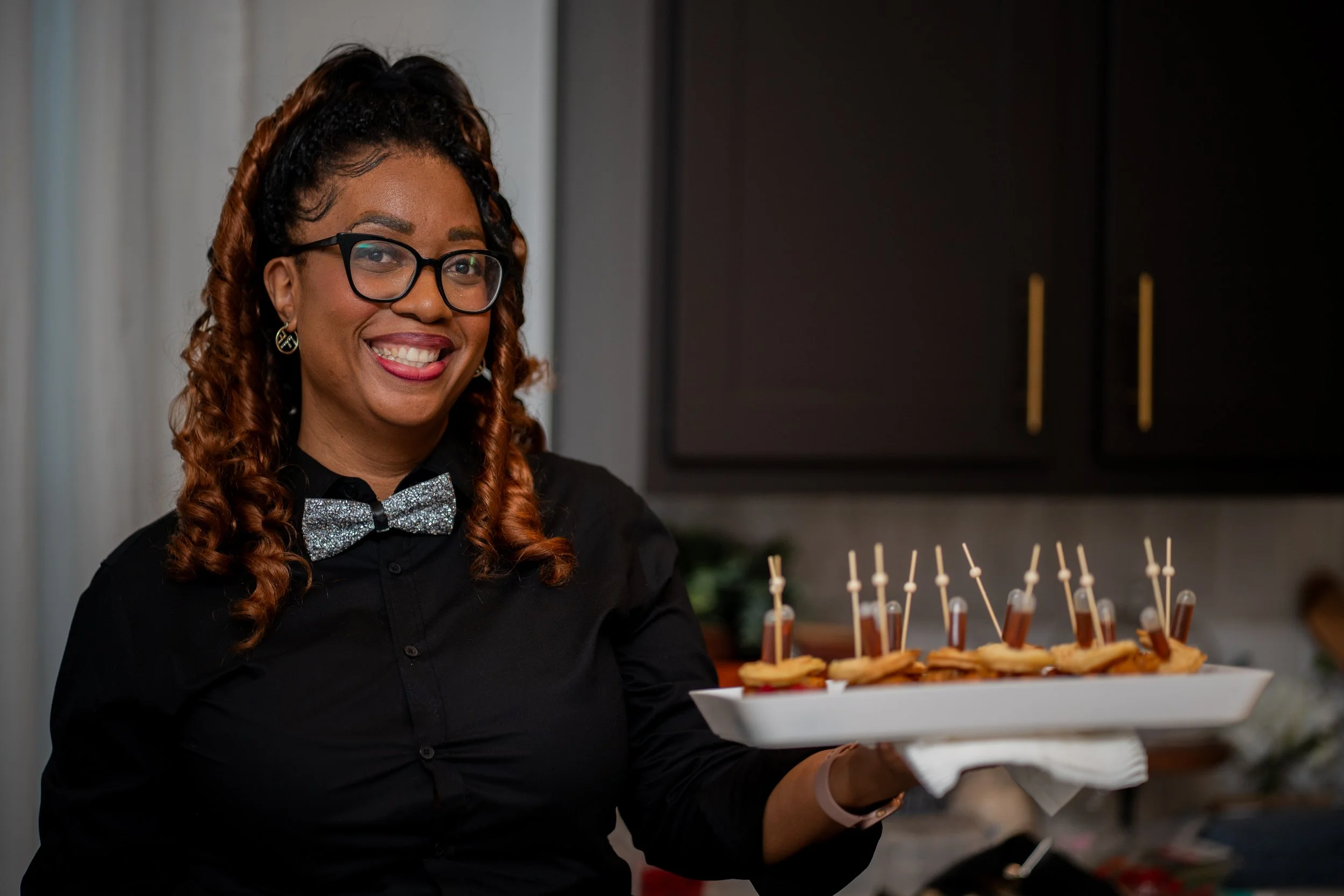 A smiling woman with glasses and curly hair holding a tray of small appetizers with toothpicks in a kitchen setting.