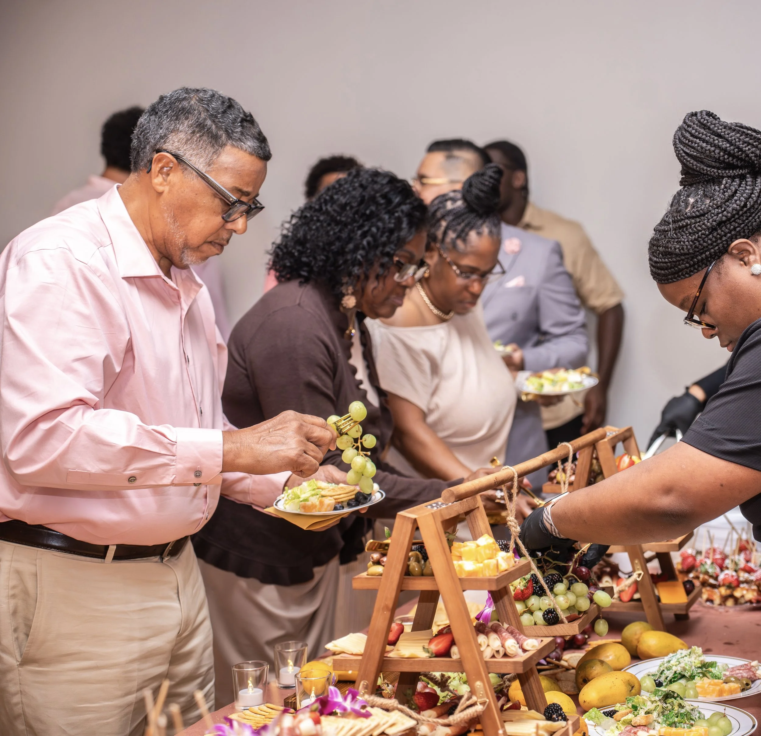 People serving themselves food at a buffet table with fruits, cheese, and snacks.