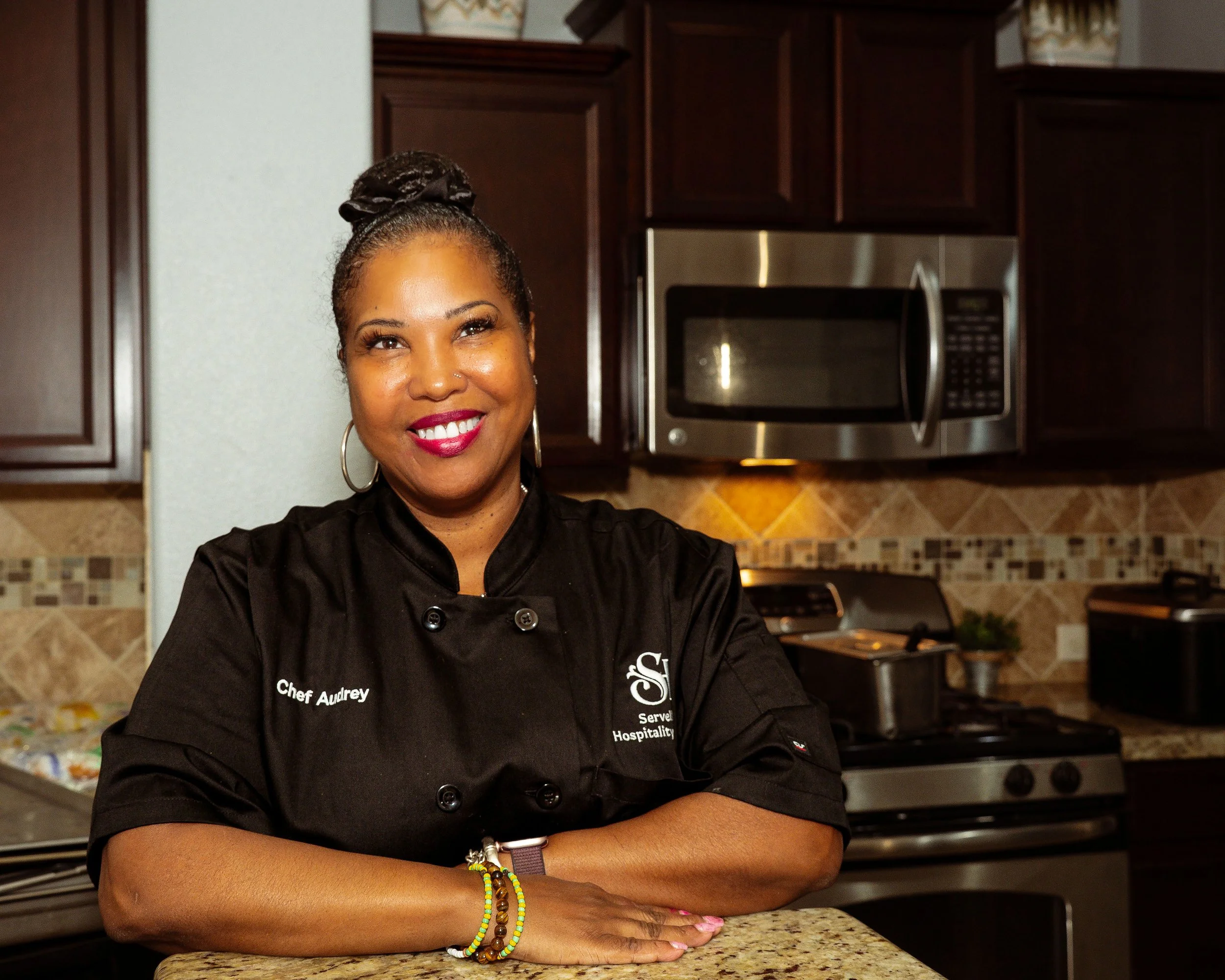 Portrait of a male chef with arms crossed, wearing a blue chef jacket, standing in a modern, softly lit restaurant or kitchen with blurred background.