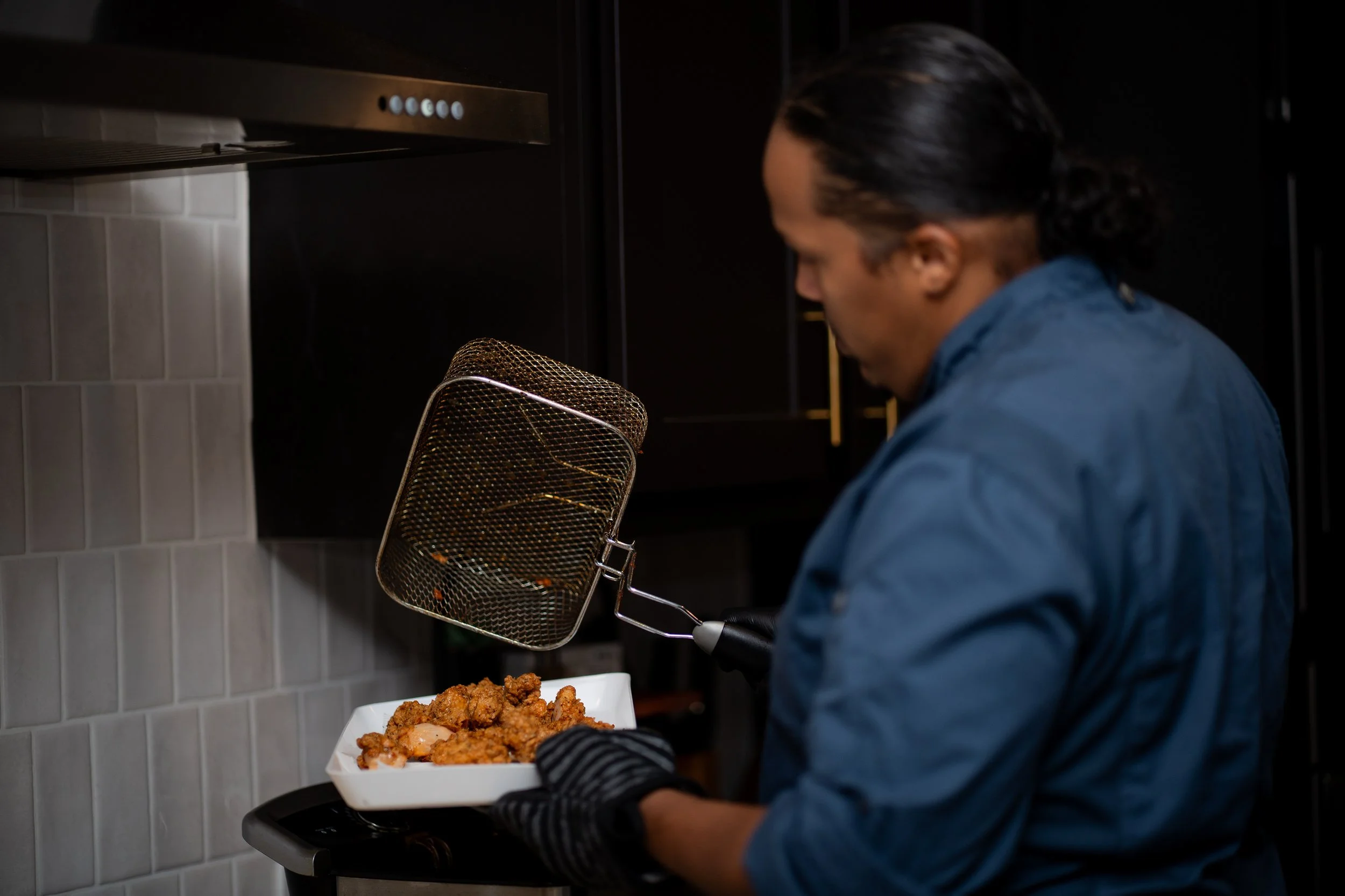 A person in a blue shirt and black gloves holding a white tray of fried chicken, with a metal fryer skimmer above it in a modern kitchen.