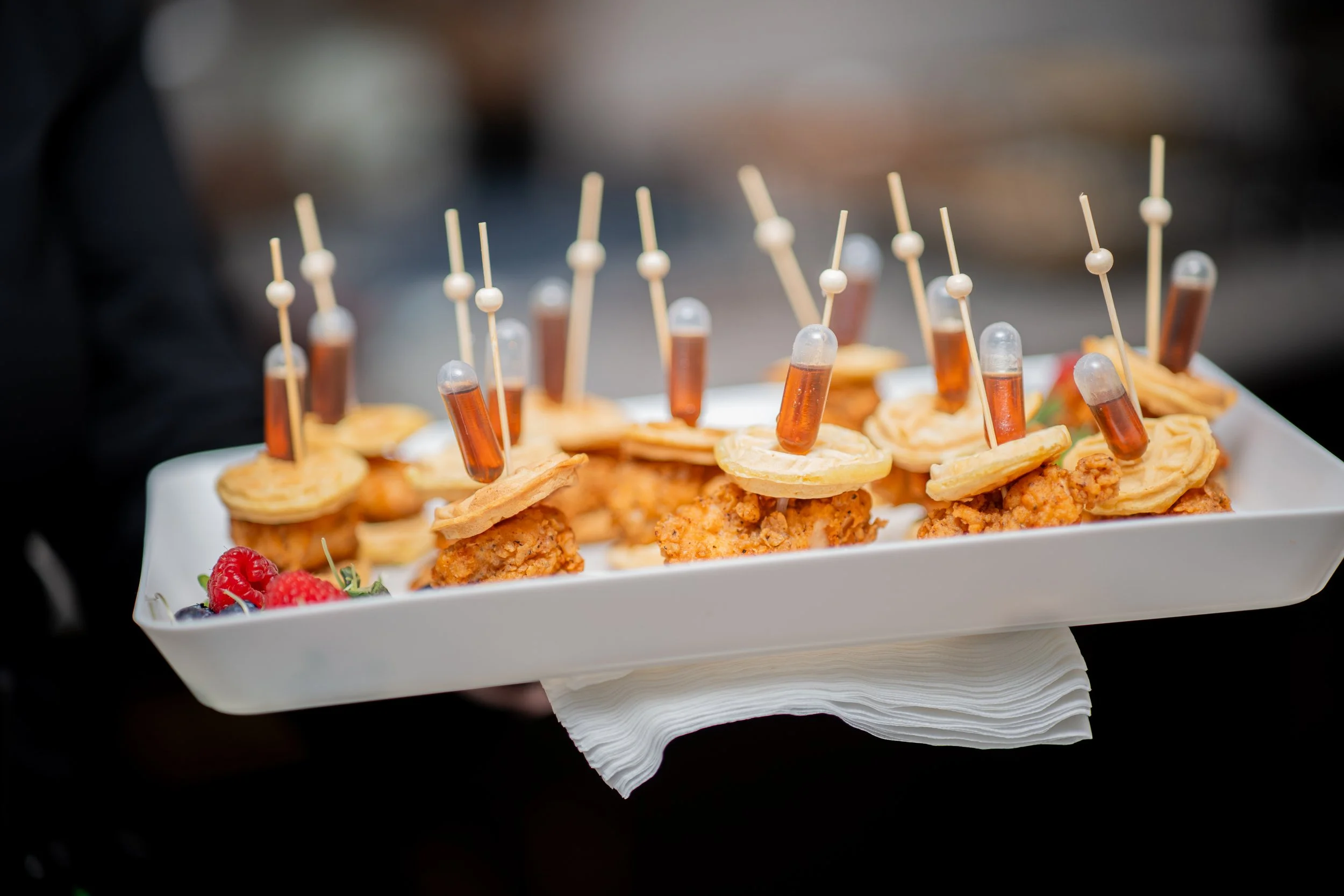 Tray with chicken bites topped with crackers, each secured with a mini syringe of sauce, garnished with berries, on a white rectangular platter.