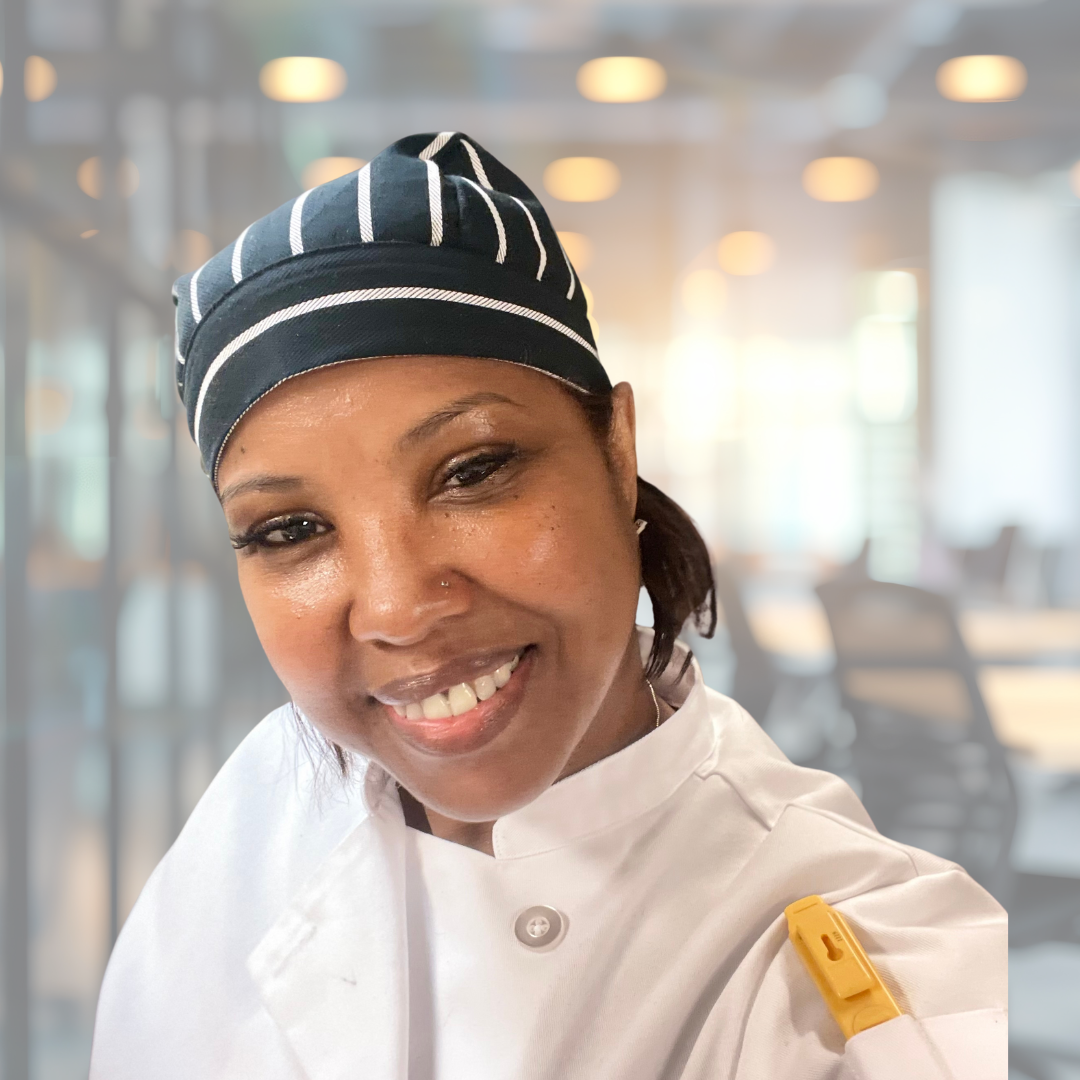 Portrait of a male chef with arms crossed, wearing a blue chef jacket, standing in a modern, softly lit restaurant or kitchen with blurred background.