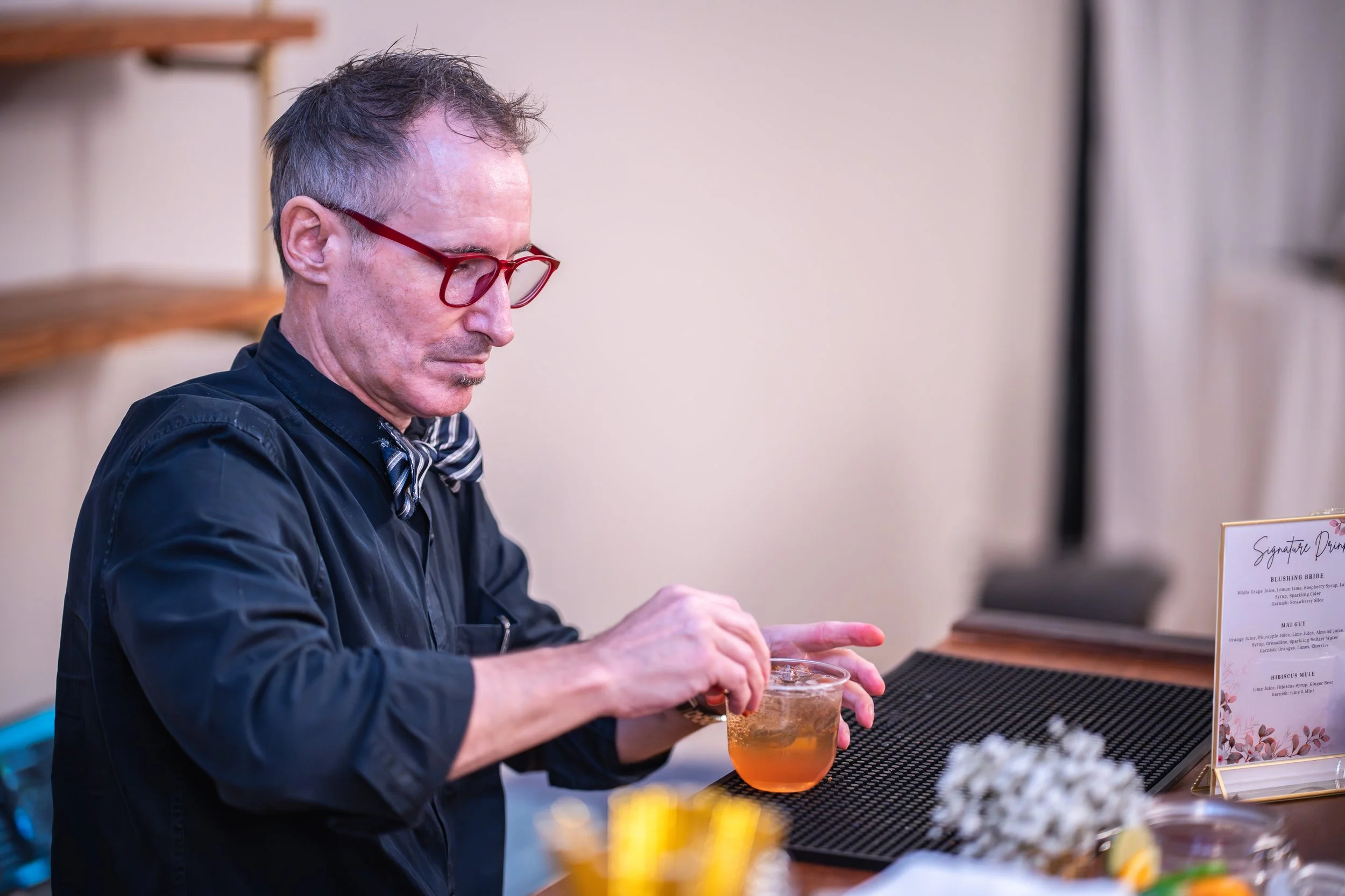 Man with red glasses and a bow tie preparing a cocktail behind a bar counter.