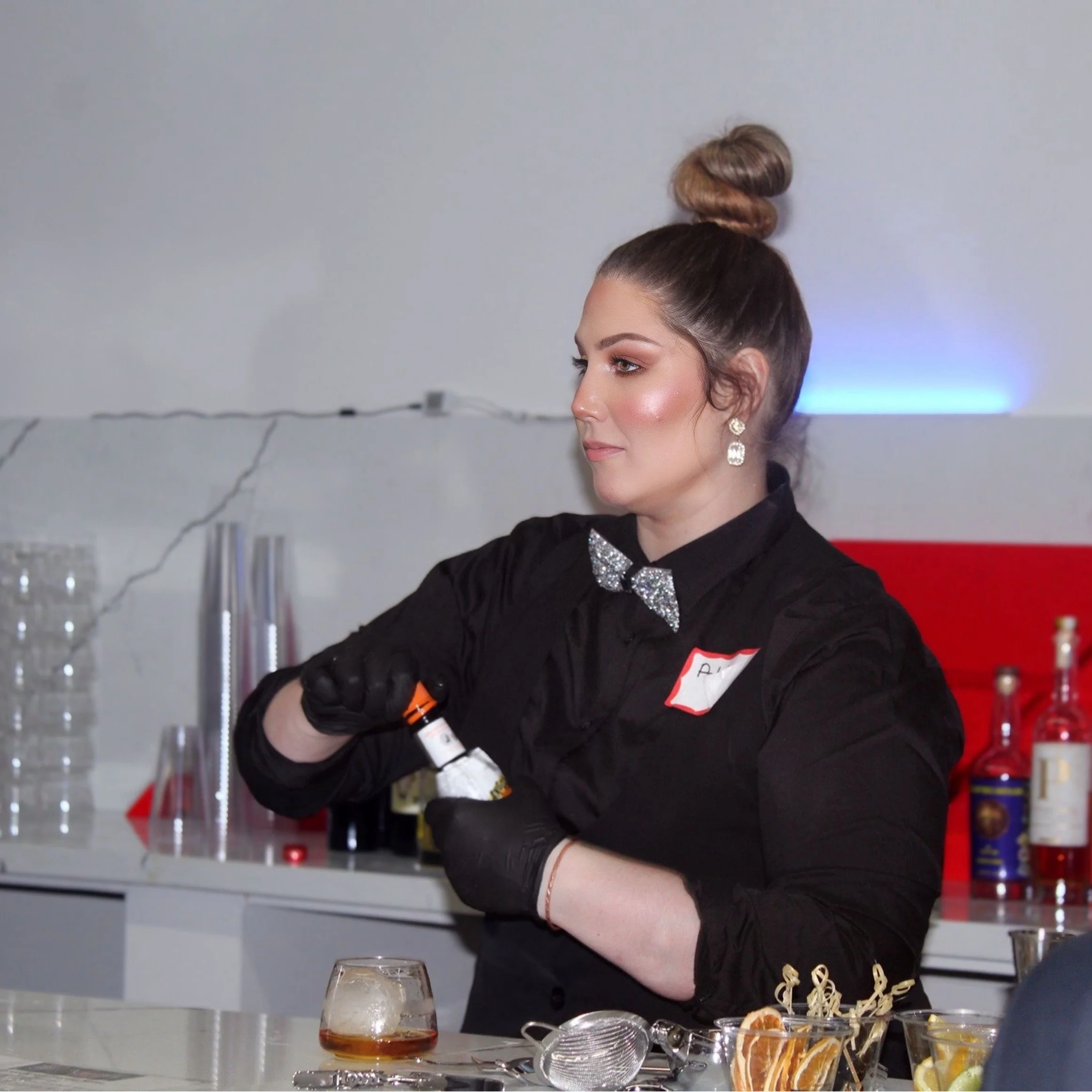 A woman with a top knot hairstyle, wearing a black shirt with a glittery bow tie and earrings, preparing a drink at a bar or event table.