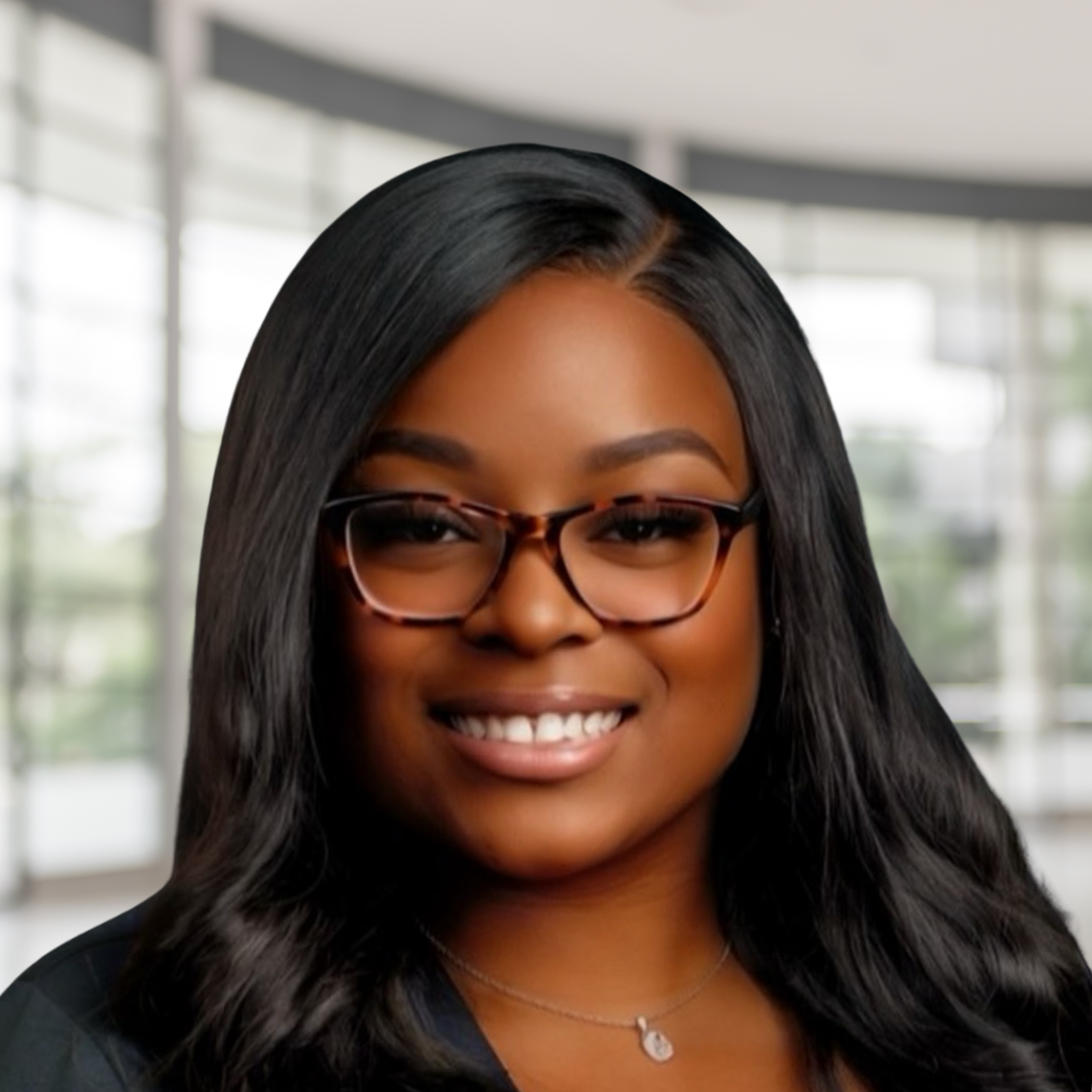 A portrait of a smiling woman with long black hair, wearing glasses, a necklace, and a dark top, with a blurred bright indoor background.