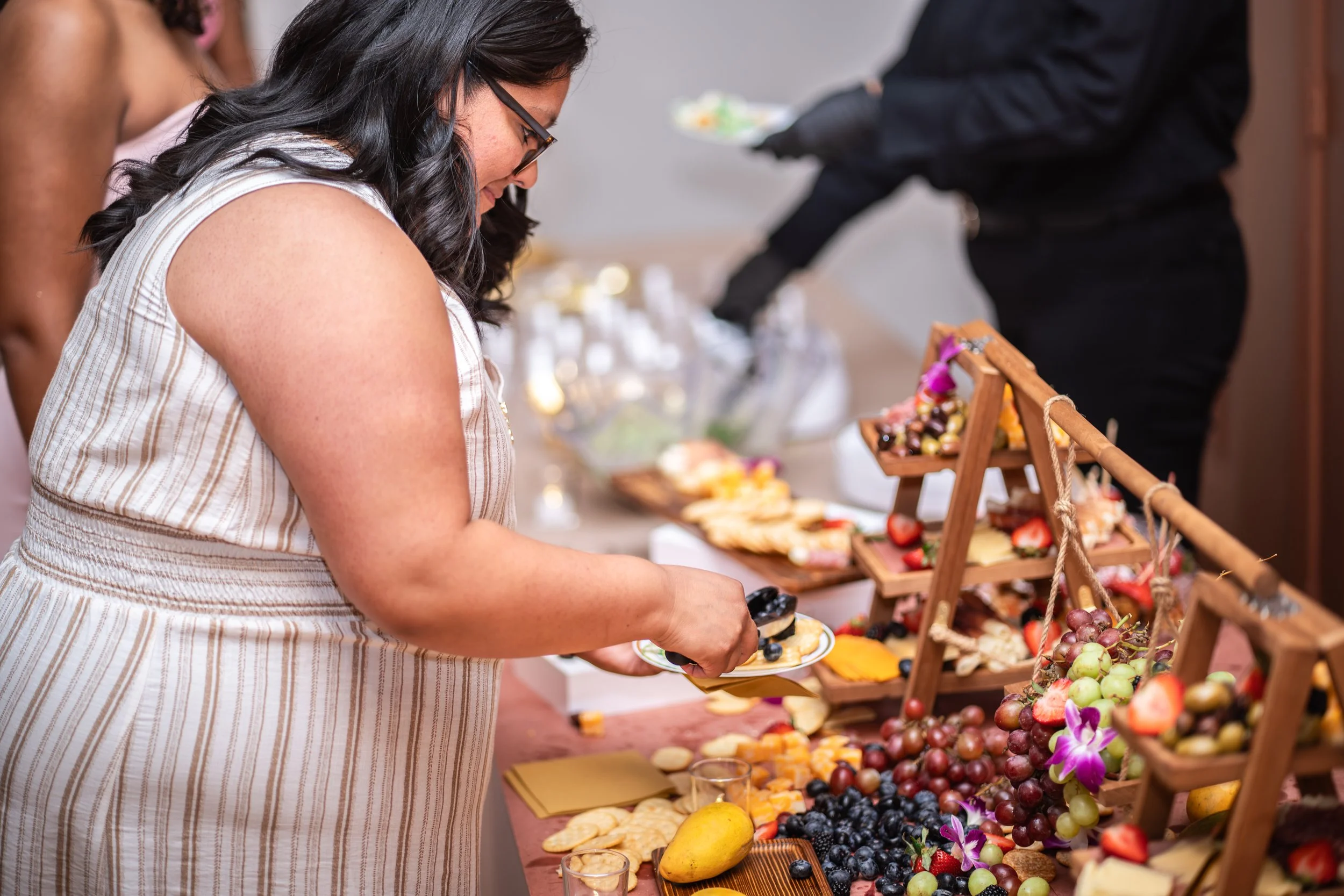 Woman in a striped dress serving herself fruit and cheese from a buffet table decorated with grapes, strawberries, blueberries, and flowers at a social gathering.