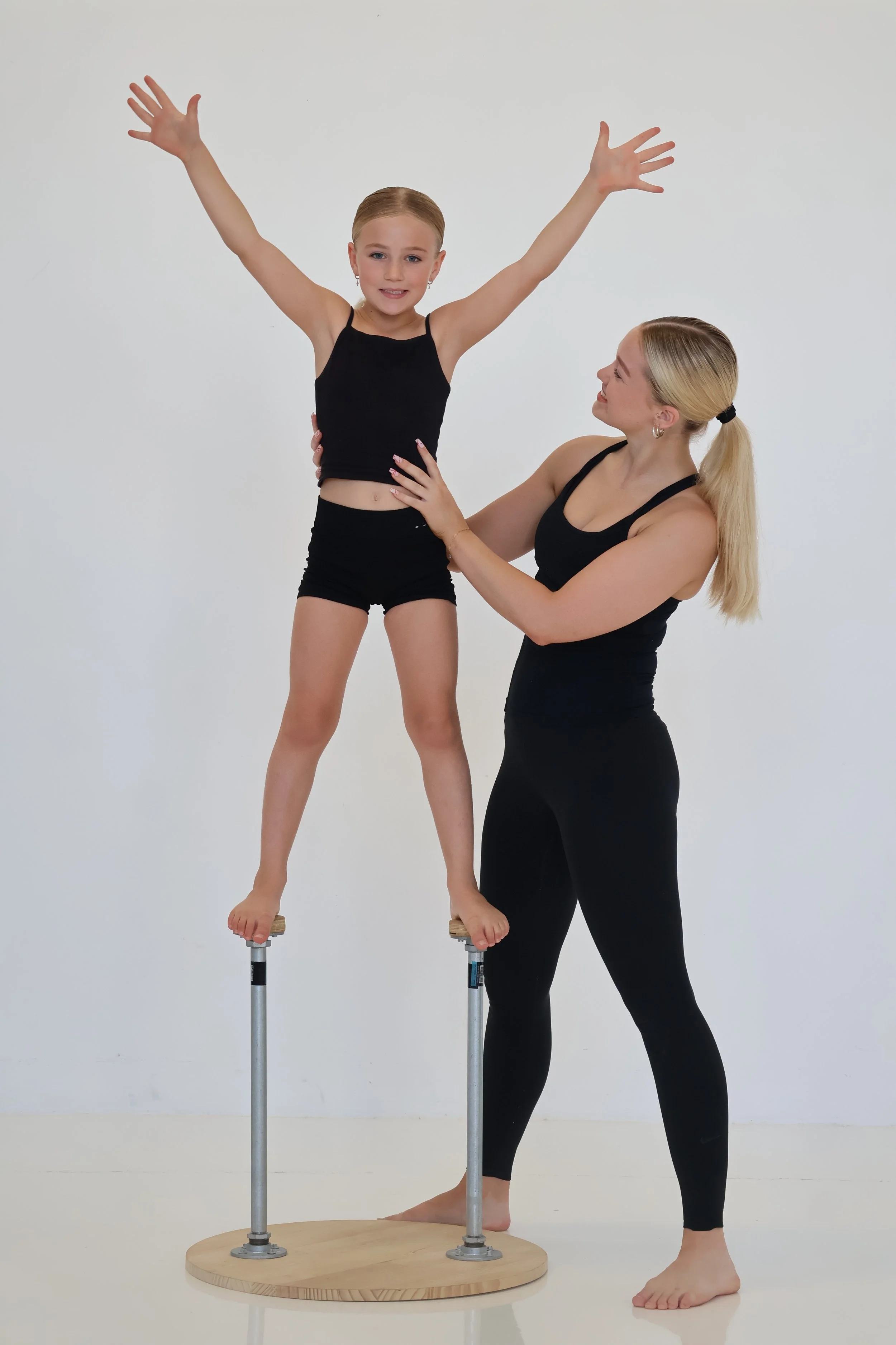 A young girl with arms raised on a balance beam, supported by a woman, against a plain white background.