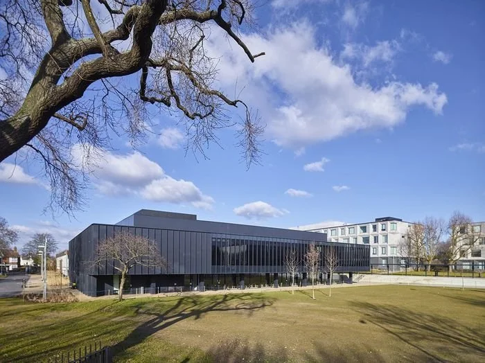 Modern black building with large glass windows, surrounded by a grassy area with few trees, under a blue sky with clouds.