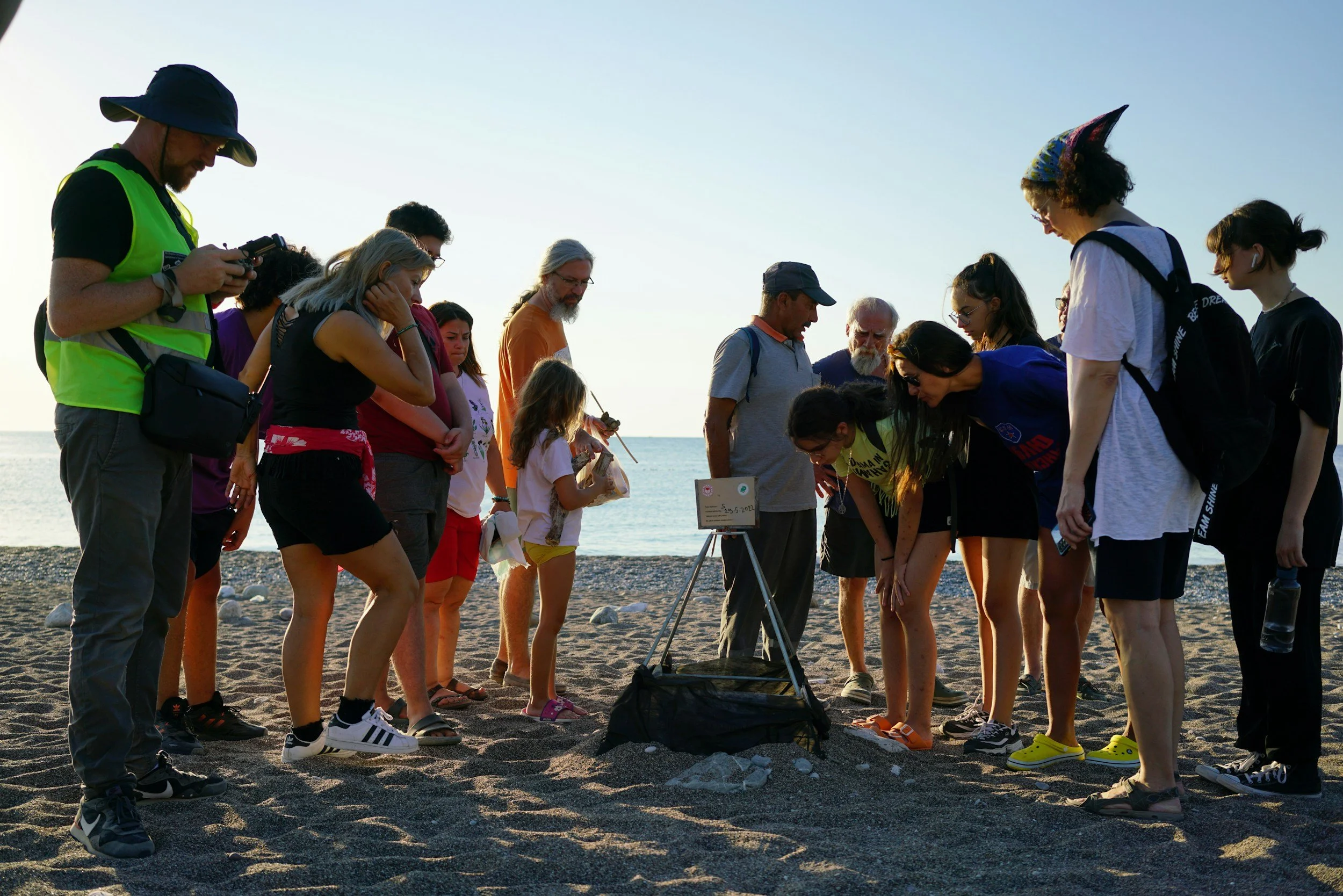 Group of diverse people gathered on a beach, looking down at something on the ground, during sunset.