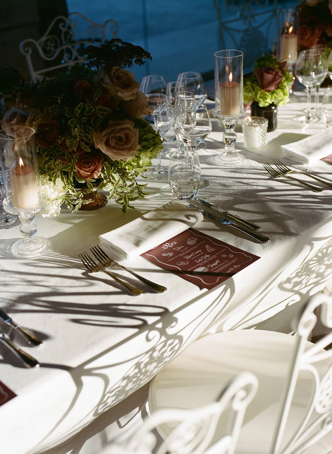 Elegant banquet table set with floral arrangements, candles, and glassware, with sunlight casting shadows.