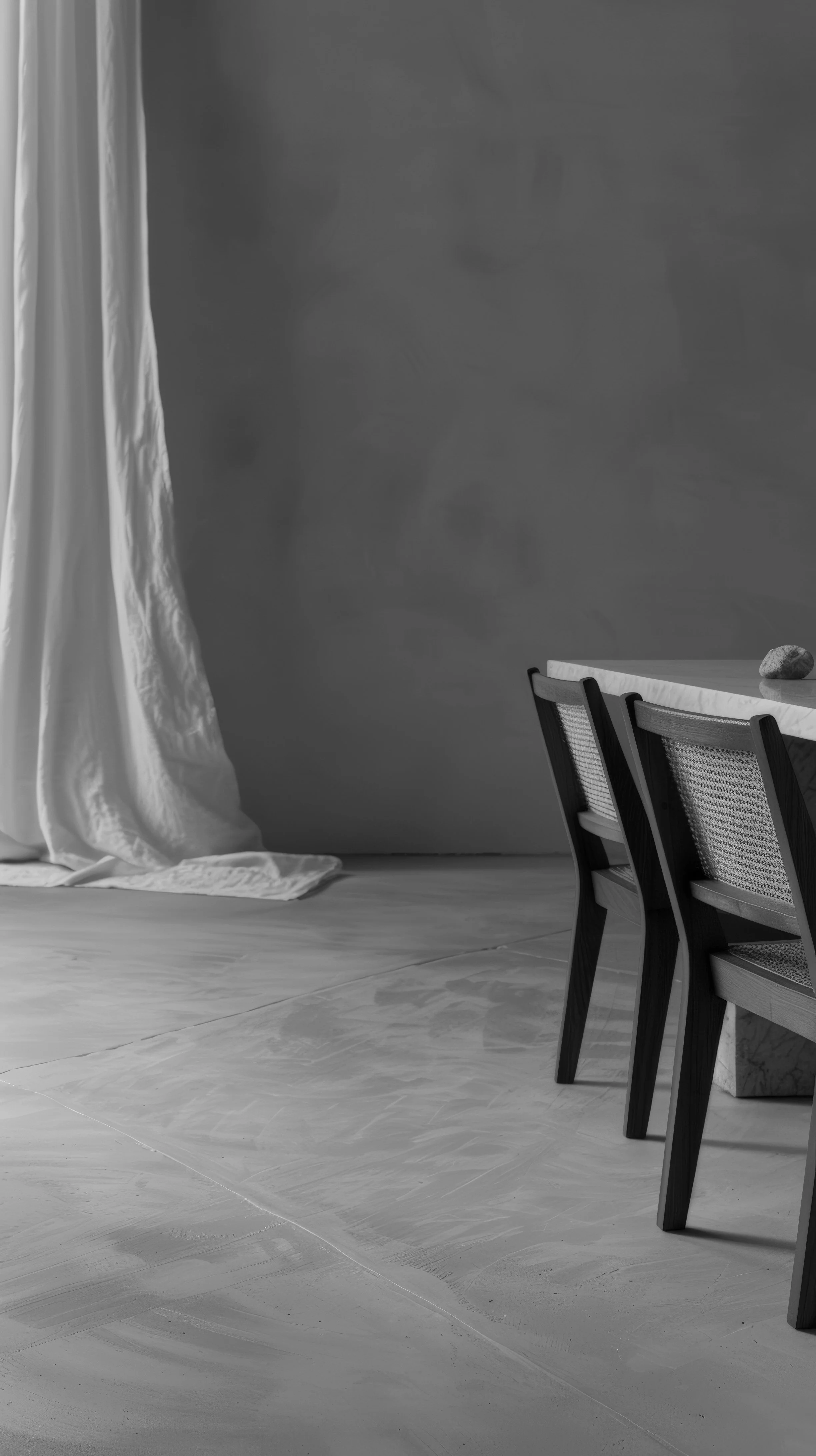 Black and white interior scene with a draped curtain on the left and a table with chairs on the right, with a small rock on the table.