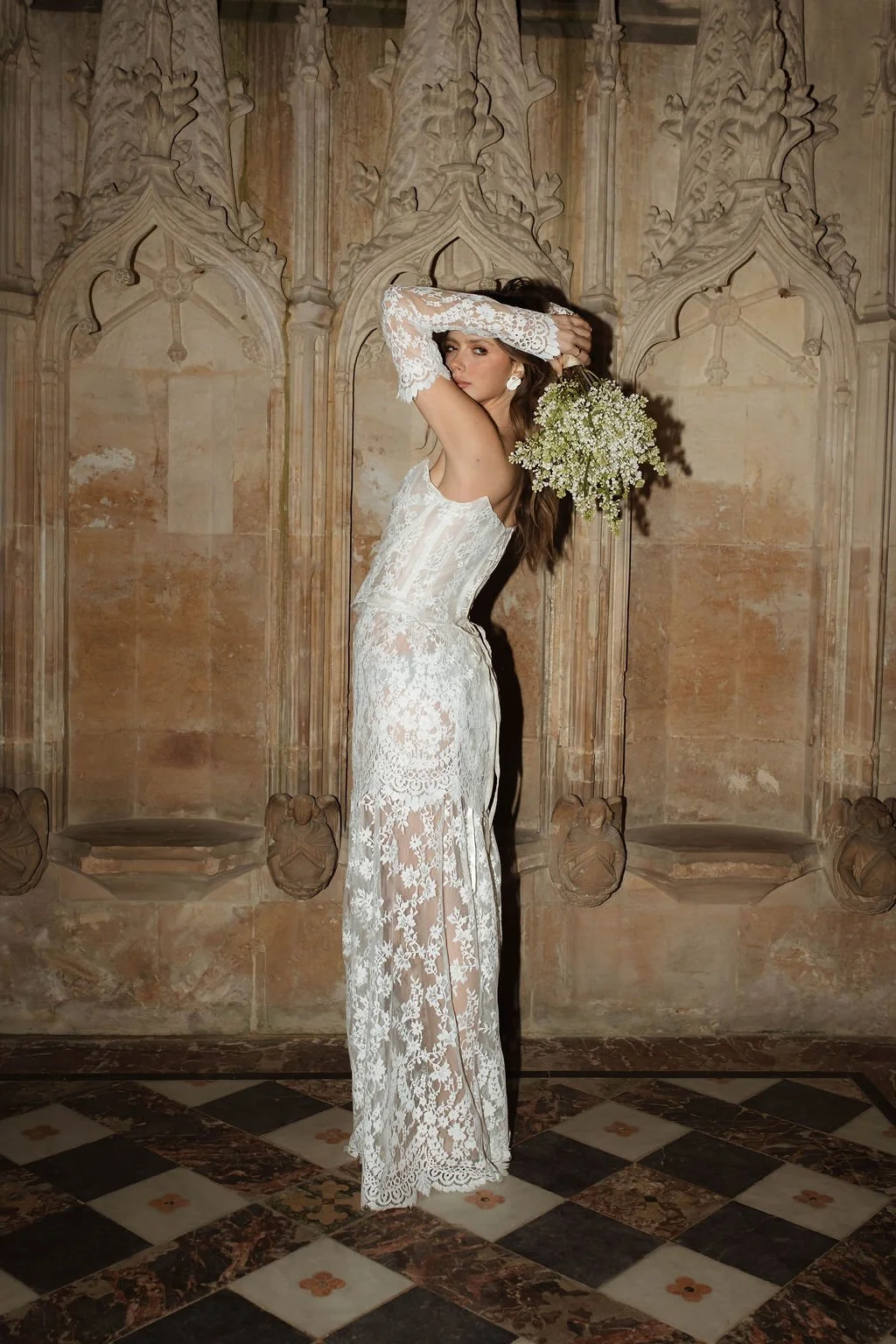 A woman in a white lace dress holding a bouquet of small white flowers standing against an ornate beige stone wall.