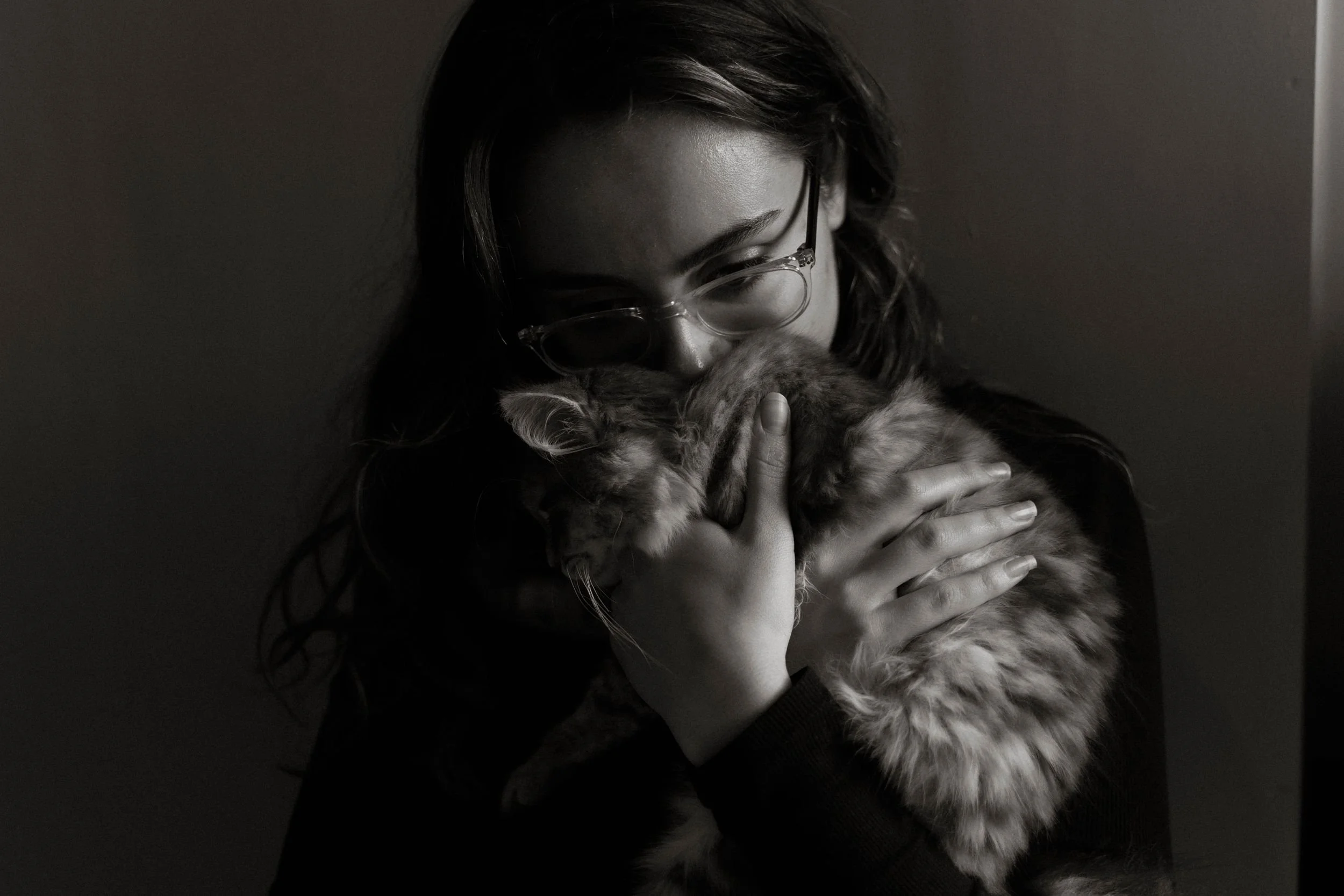 A woman with glasses holding and cuddling a fluffy, long-haired cat in a dimly lit room.