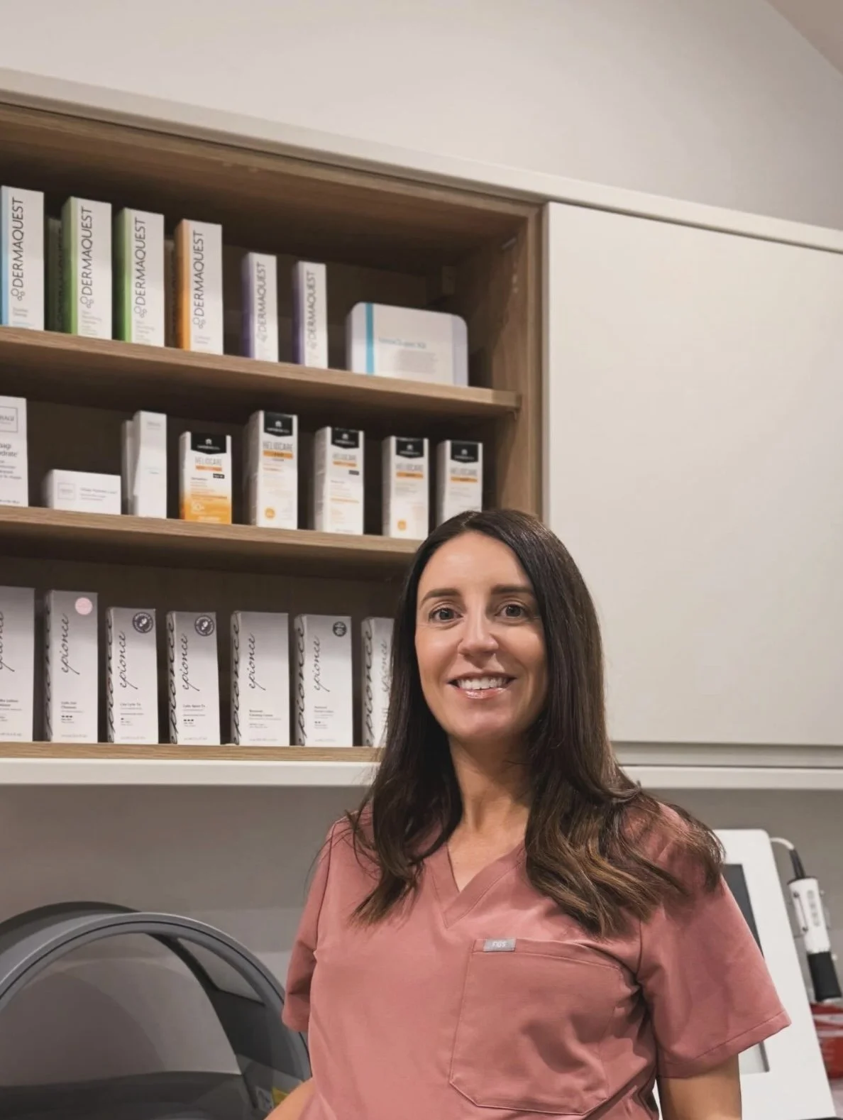 A woman in medical scrubs standing in front of shelves filled with skincare products in a clinic or pharmacy.