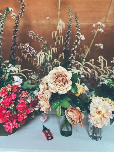 Arranged flowers including roses, geraniums, and various wildflowers in vases, with gardening scissors on a table.