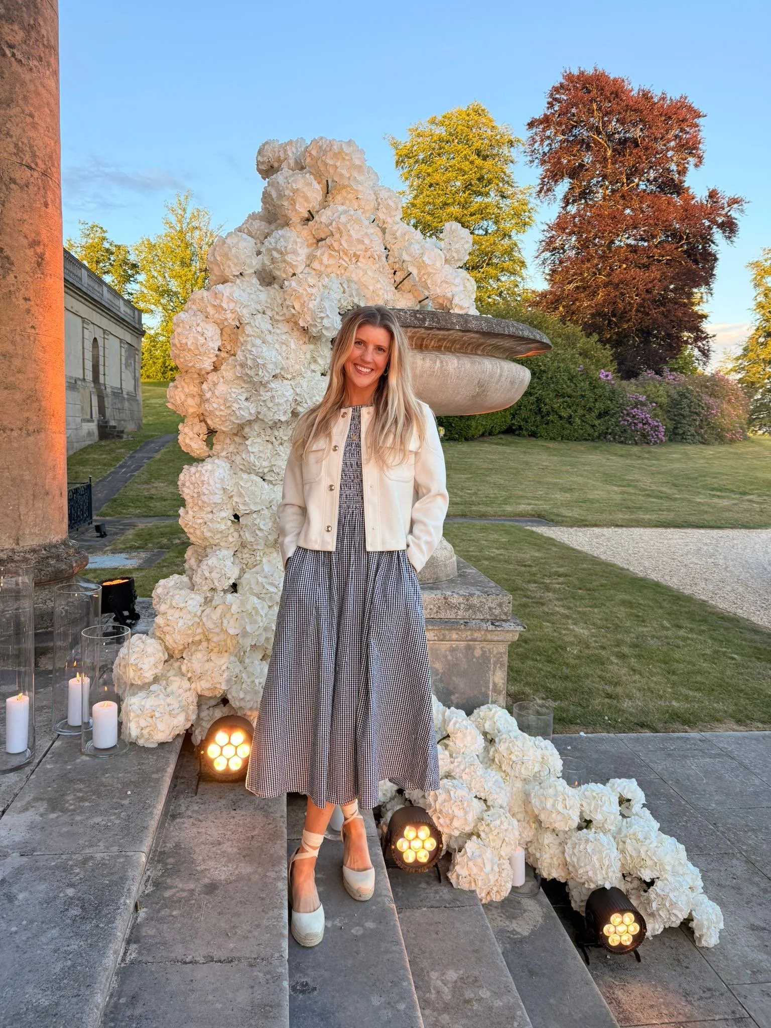 A woman in a checkered dress, white jacket, and white high heels is standing in front of a floral decorative display with white hydrangeas and candles, outdoors during sunset with trees in the background.