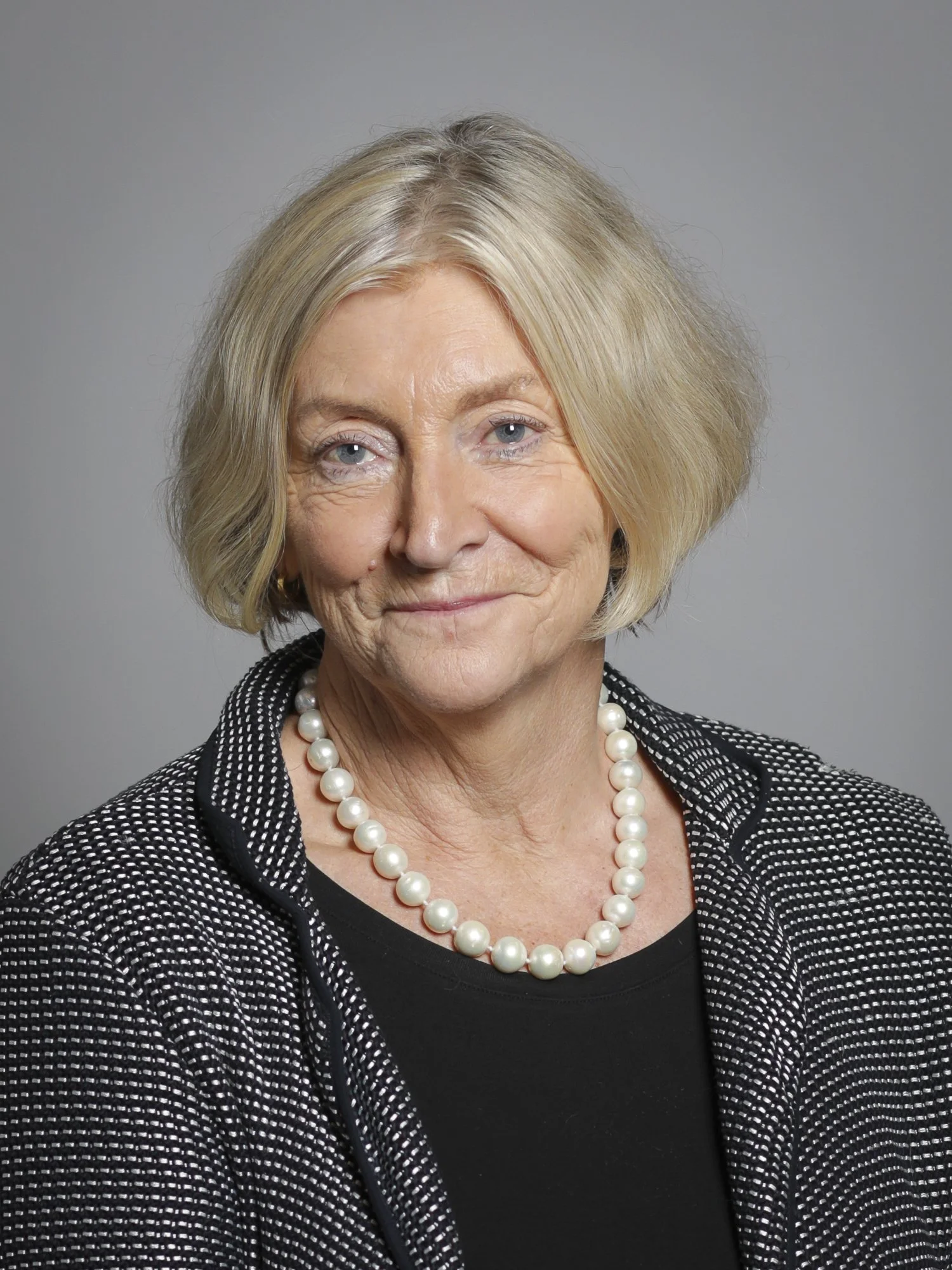 A portrait of an elderly woman with short blonde hair, blue eyes, and a gentle smile. She is wearing a black top, a pearl necklace, and a black and white patterned jacket against a plain gray background.