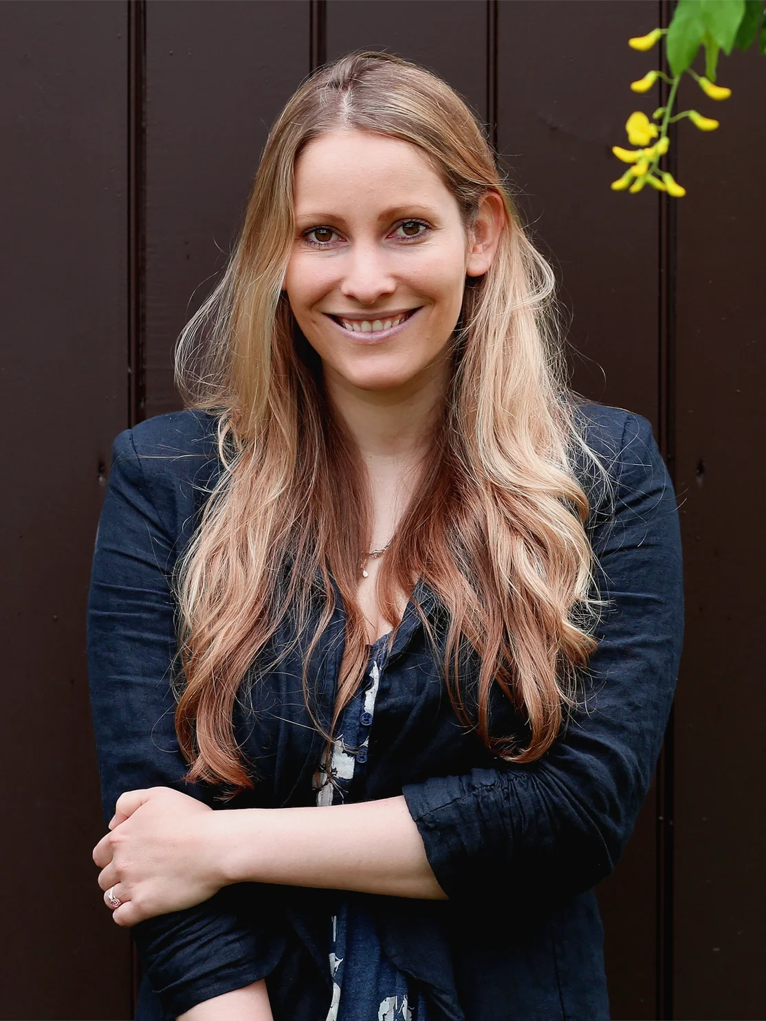 A smiling young woman with long wavy blonde hair, wearing a dark blue jacket, standing outdoors in front of a dark wooden fence, with some green leaves visible in the upper right corner.