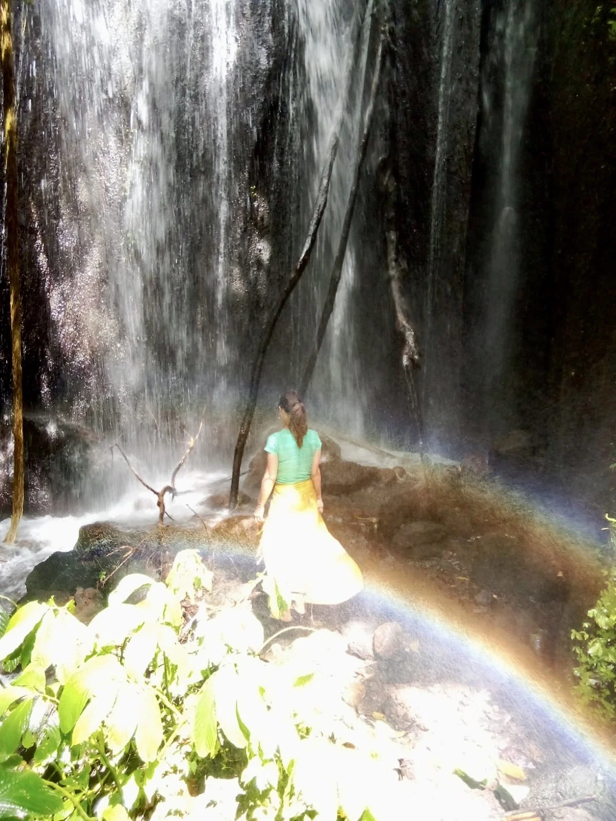 A woman standing on rocks at the base of a waterfall, with a rainbow appearing at the bottom of the waterfall, surrounded by lush greenery.