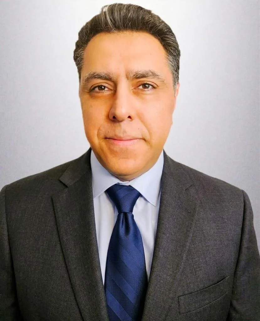 Headshot of a man with dark hair wearing a gray suit, a light blue shirt, and a dark blue tie, standing against a plain white background.
