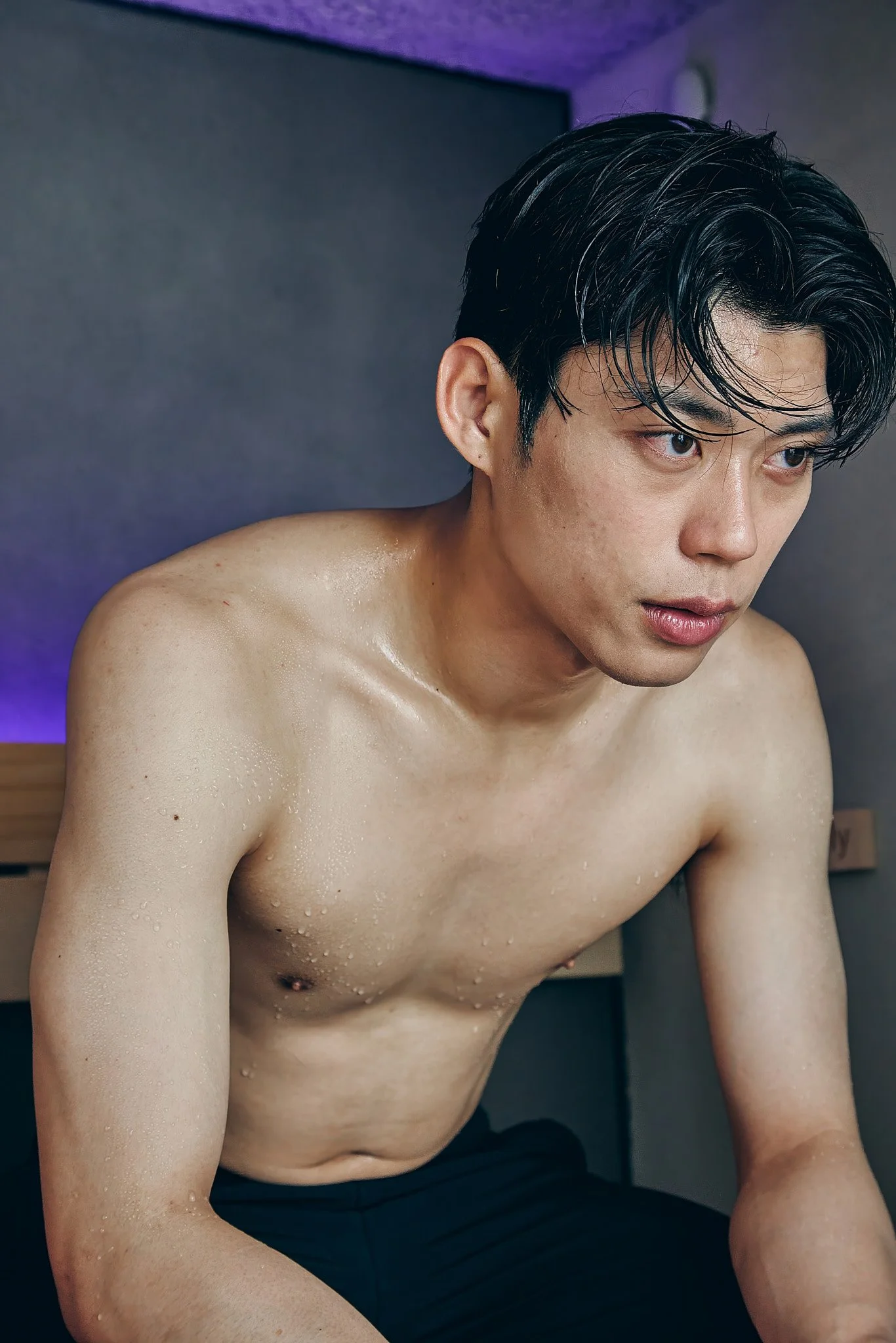 A young Asian man with wet hair and shirtless, looking thoughtful, sitting indoors.