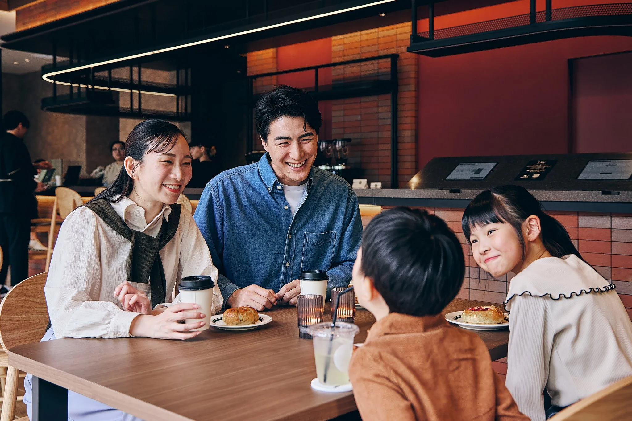A group of four people, two adults and two children, sit at a table in a cafe, smiling and enjoying drinks and pastries.