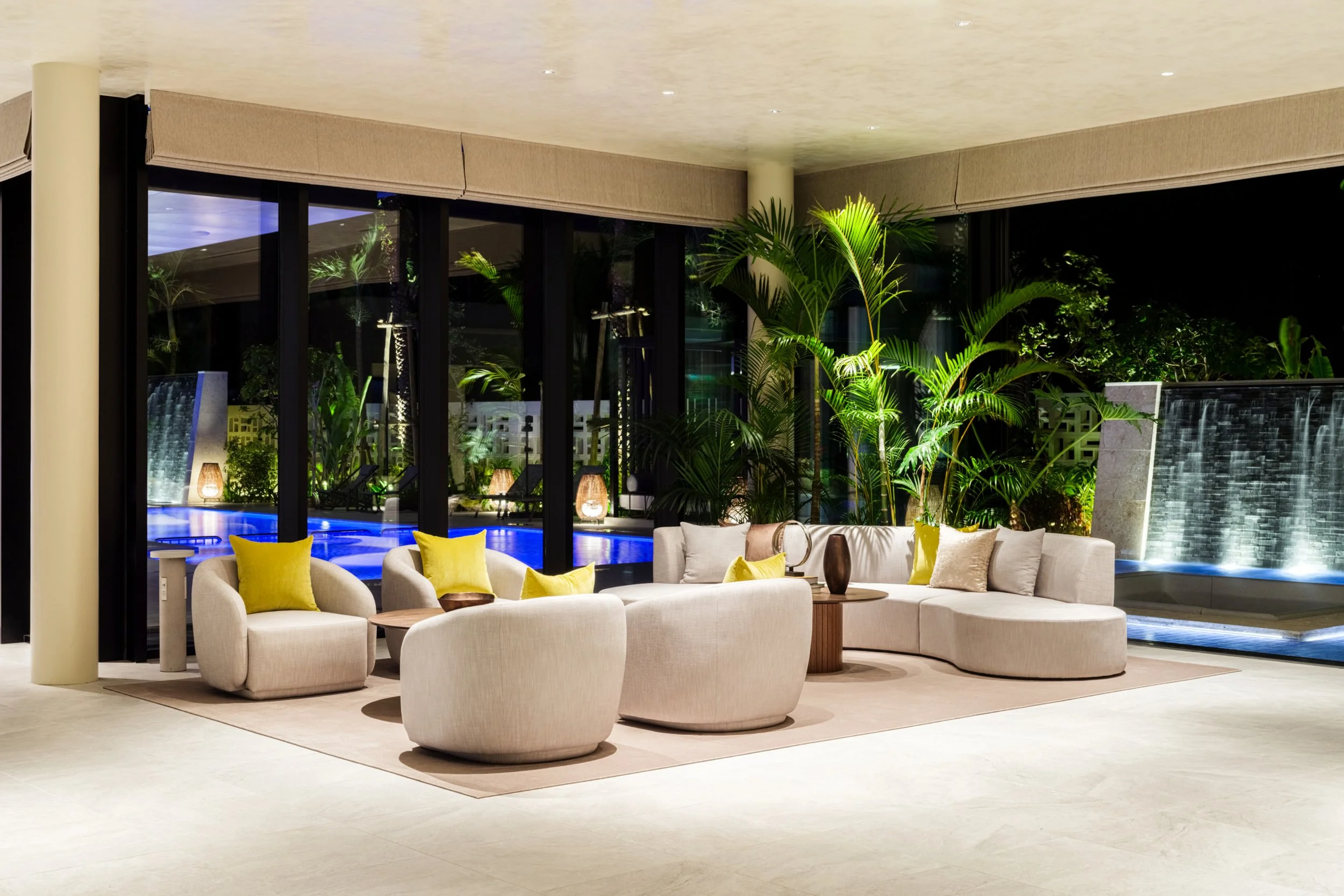 Luxury hotel lobby with modern beige seating and large green potted plants, overlooking an outdoor pool area through floor-to-ceiling glass windows at night.