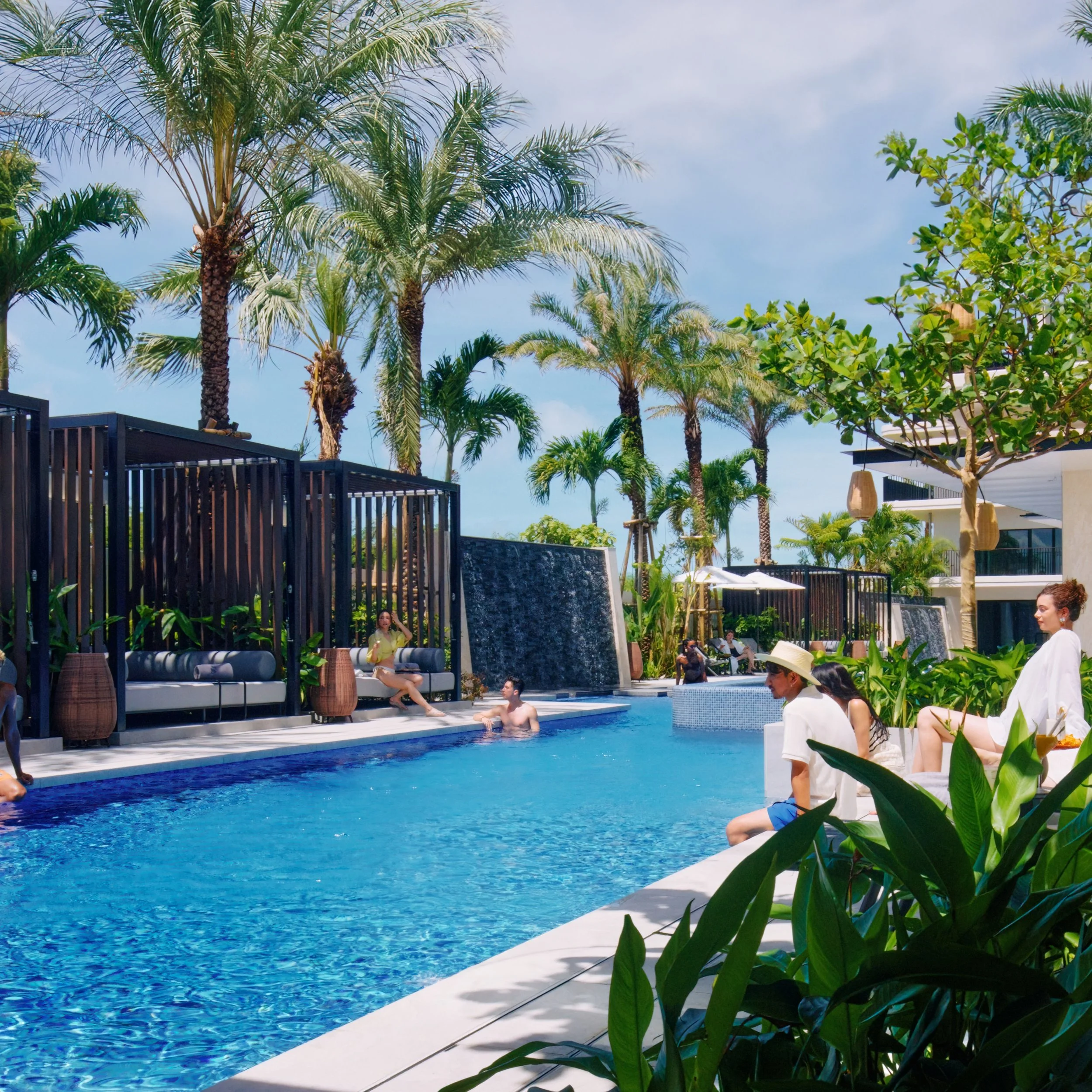 People relaxing by a pool surrounded by palm trees and tropical plants at a resort with modern architecture.