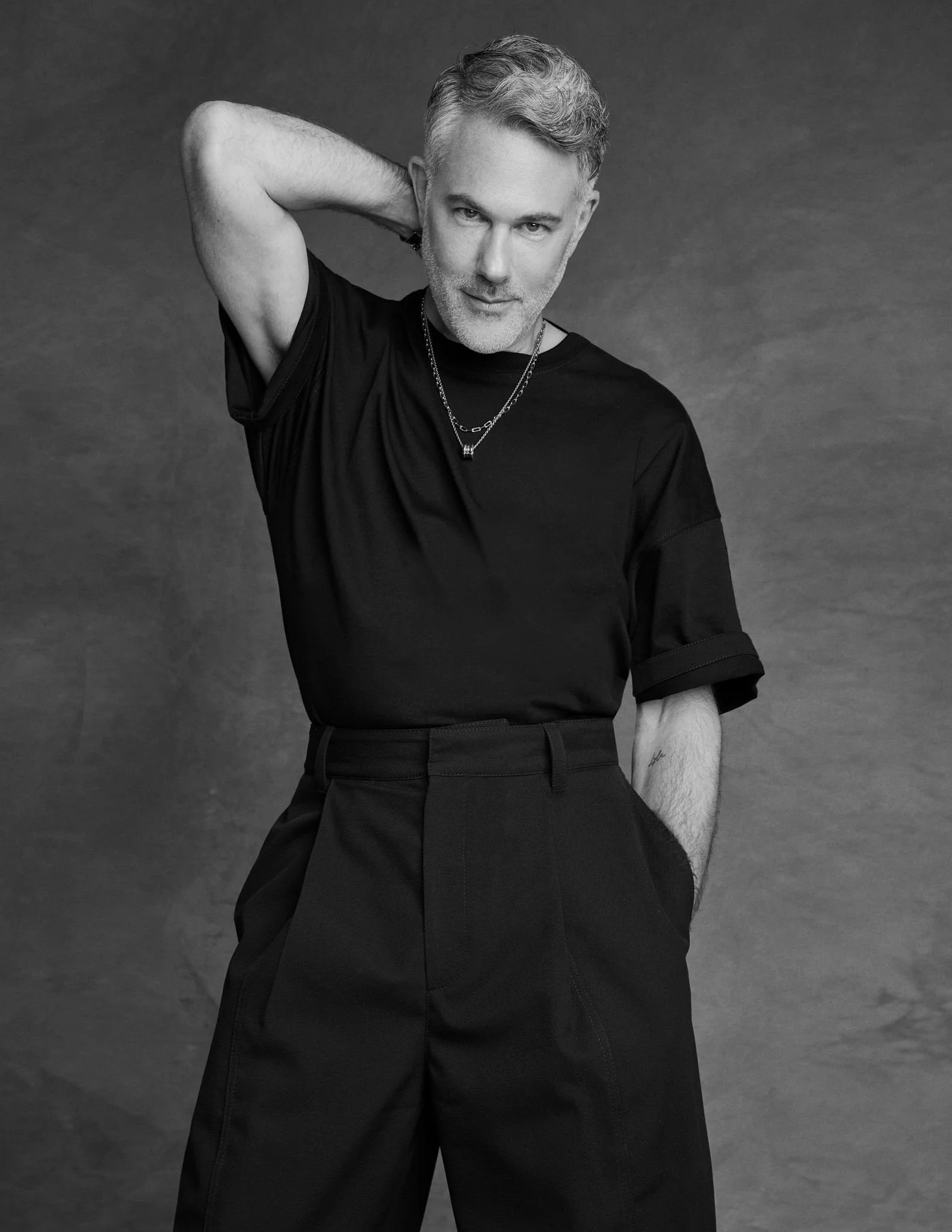 Black and white photo David Miskin with short, gray hair wearing a black t-shirt, dark pants, a necklace, and with one hand behind his head, looking at the camera.