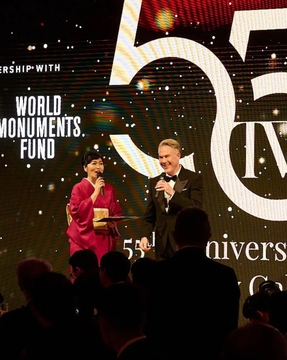 A woman in traditional Japanese attire and a man in a tuxedo (David Miskin) hosting the 55th anniversary event, with a large LED screen in the background displaying the logo for the 55th anniversary of the World Monuments Fund.