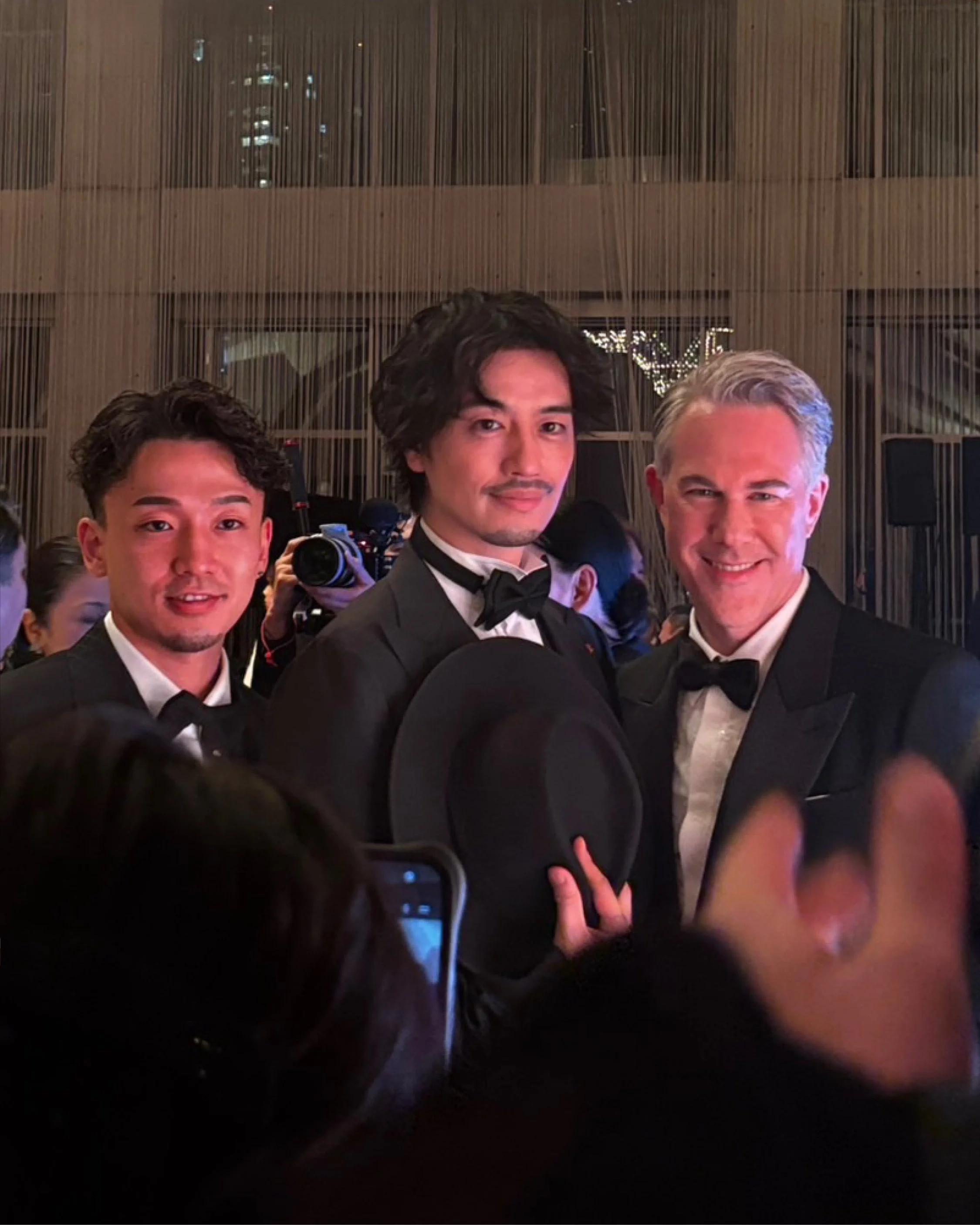 Three men in tuxedos at a formal event, one holding a black hat with a wide brim, surrounded by a crowd and photographers.