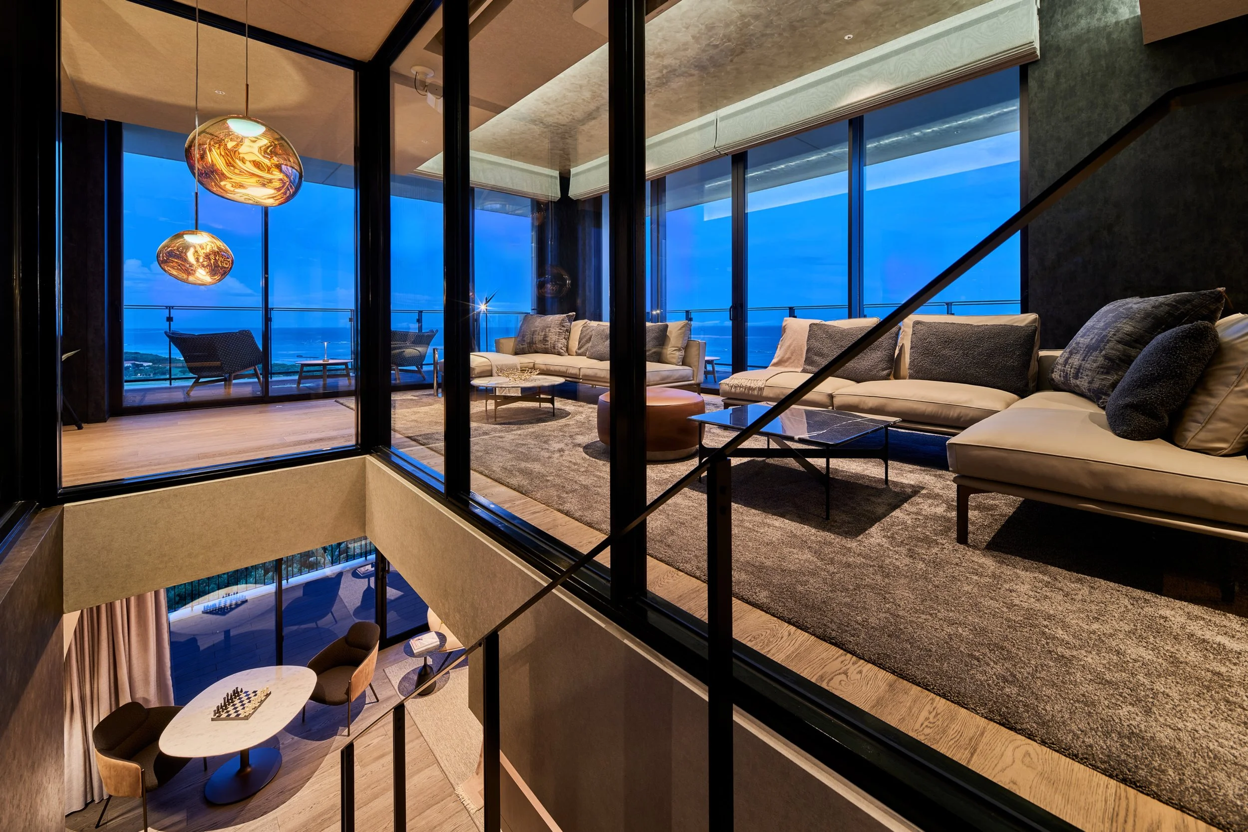 Modern living room with large floor-to-ceiling windows offering a view of the city or ocean, furnished with beige sofas, throw pillows, and a coffee table, illuminated by pendant lights, with an adjacent dining area below.