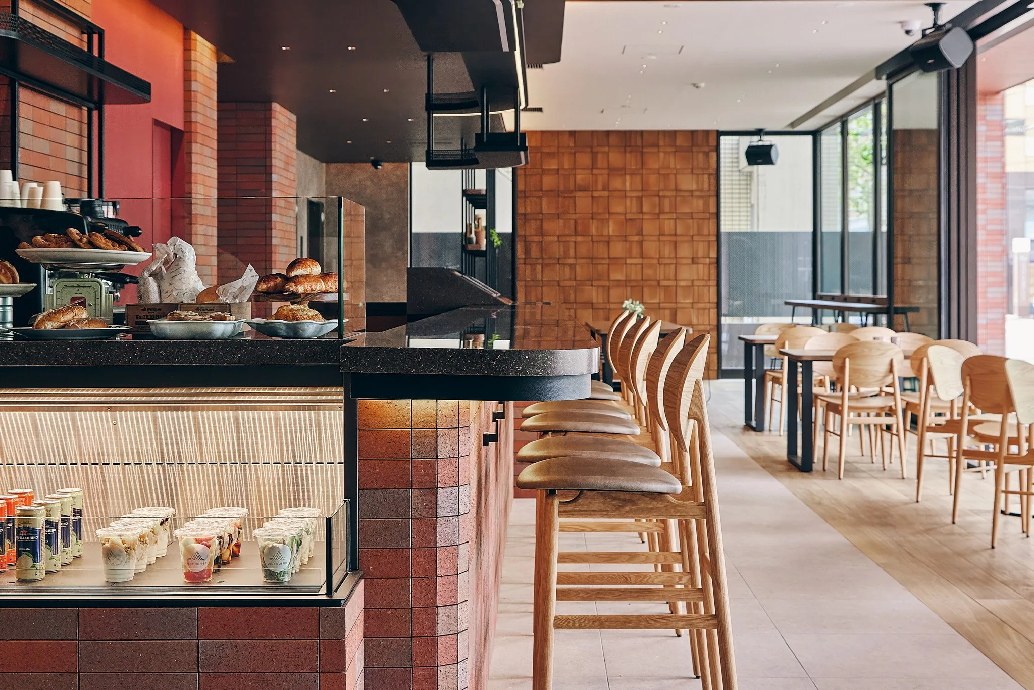 An empty cafe with wooden chairs and tables, a counter with baked goods and yogurt cups, and large windows letting in natural light.