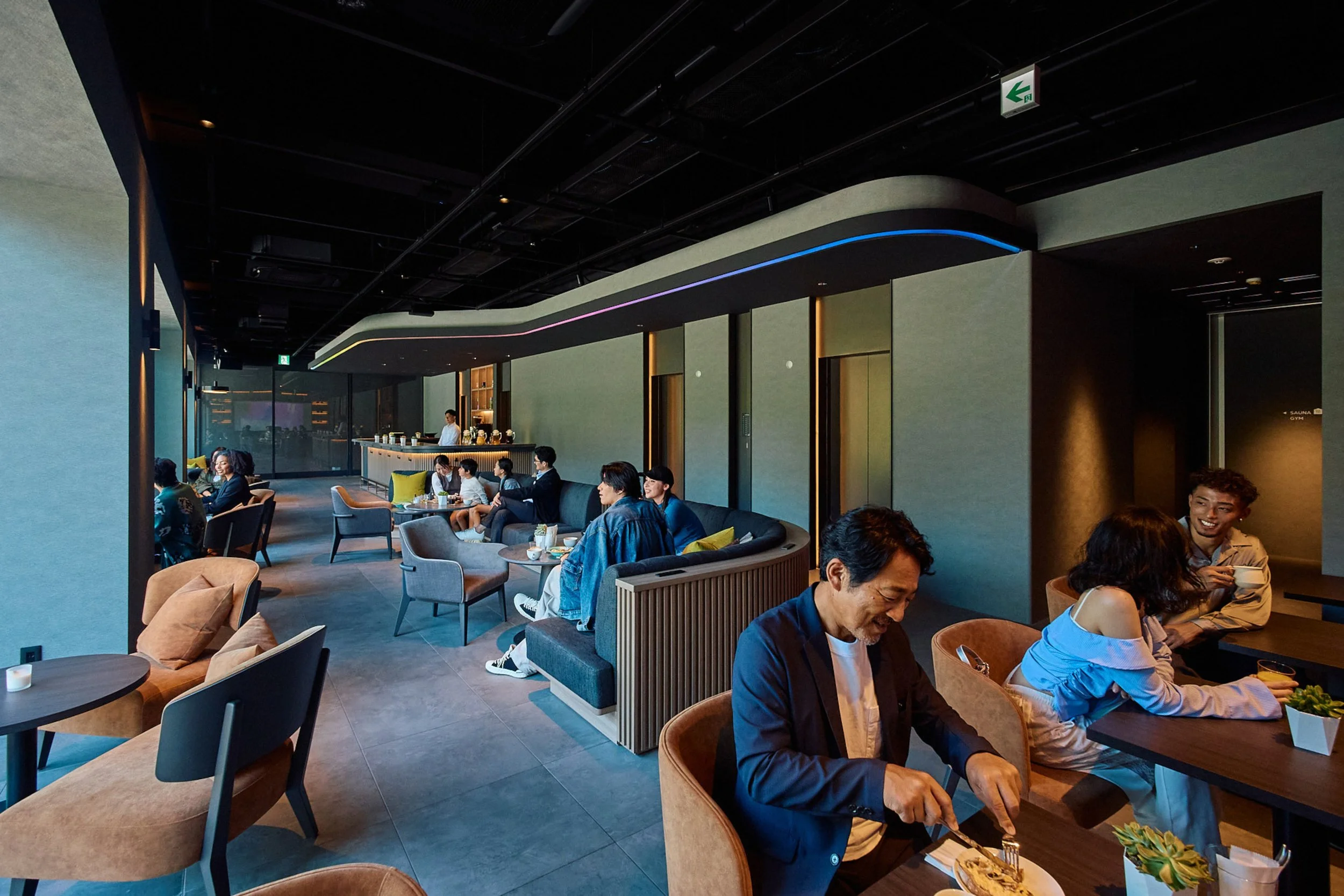 People dining and socializing in a modern, stylish restaurant with a bar area in the background and colorful LED lighting on the ceiling.