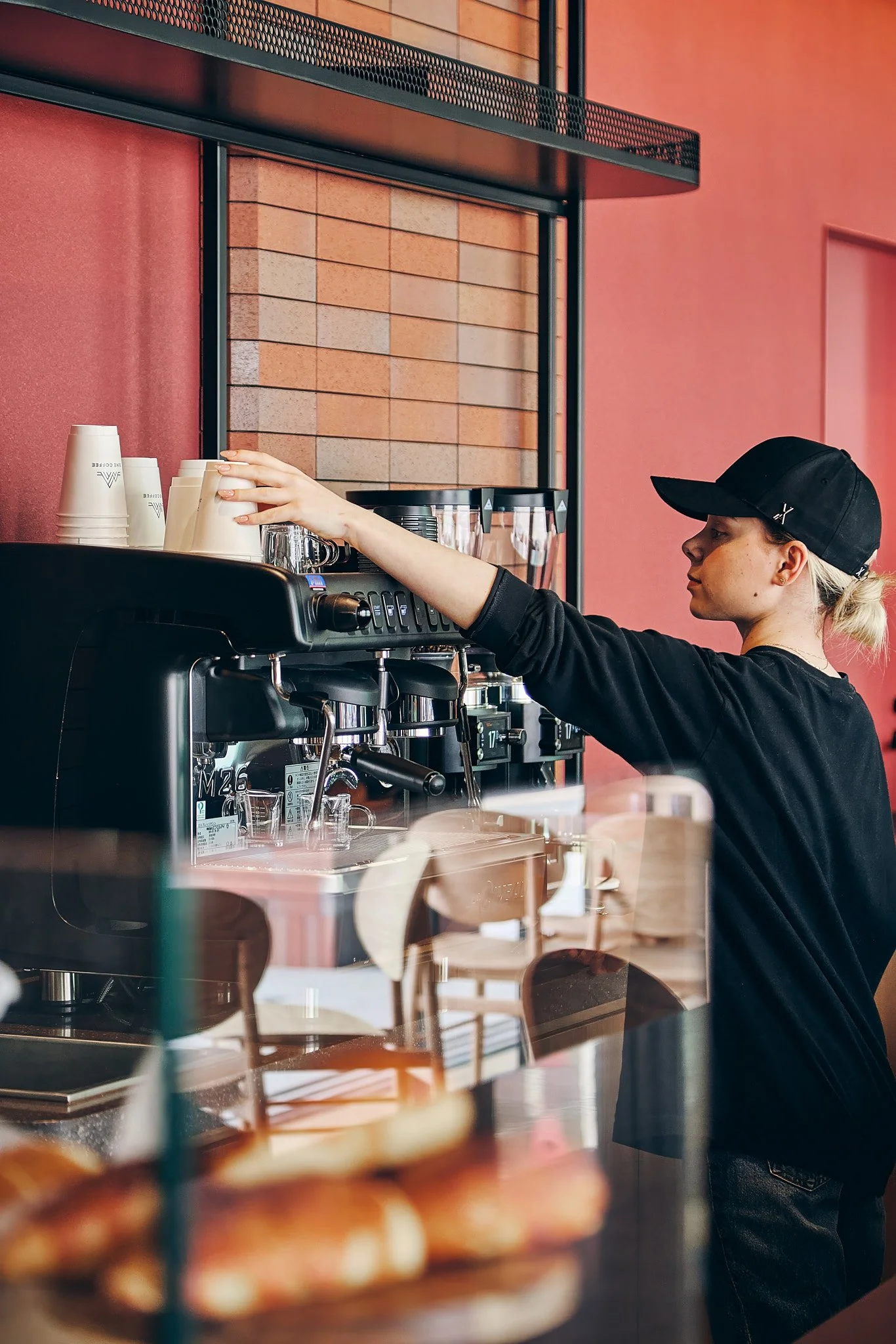 A barista wearing a black cap and black shirt operating an espresso machine behind a coffee bar, with cups stacked on top and baked goods in the foreground.