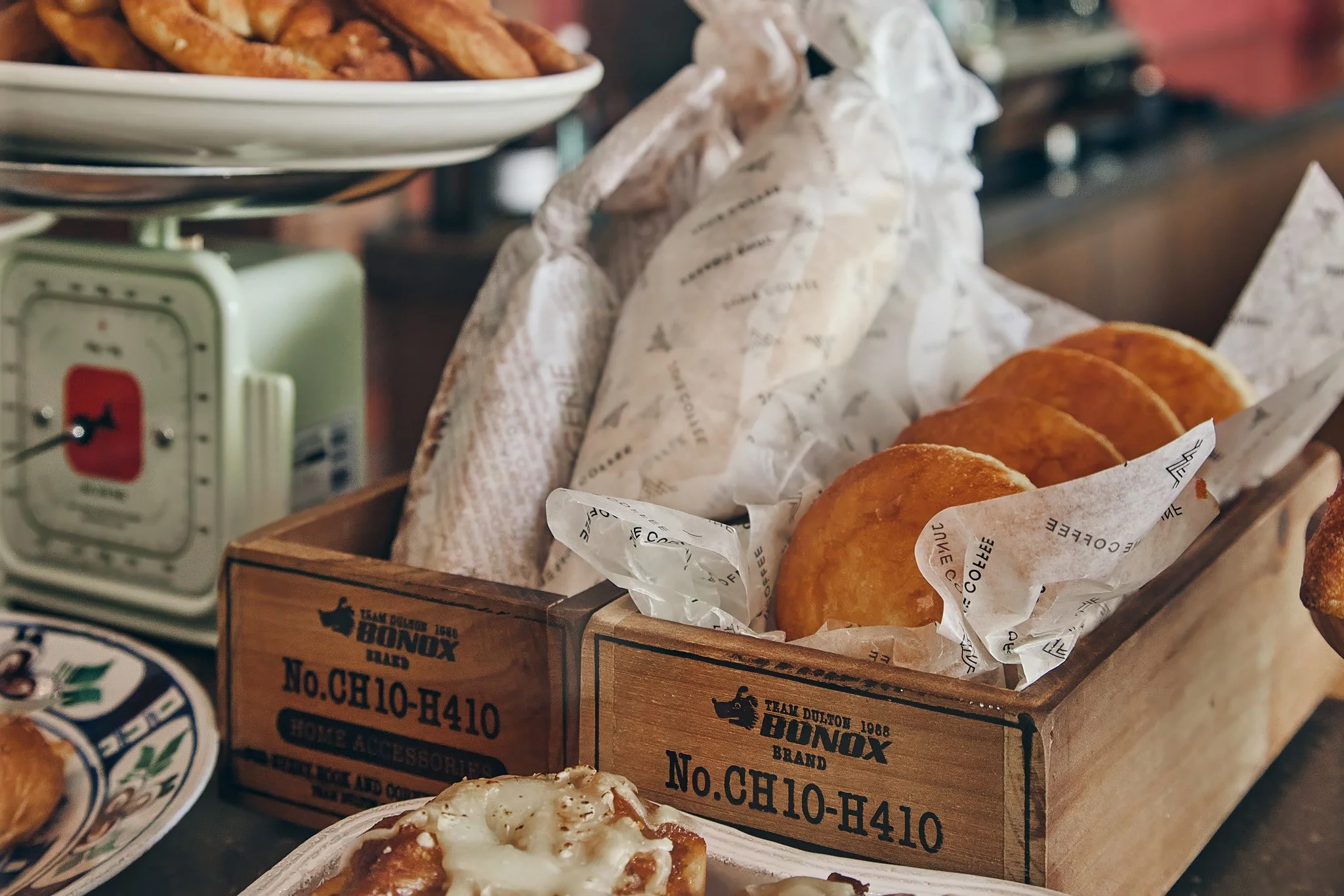 A wooden box filled with fried eggs with a paper liner, with a plate of pizza and a vintage green scale in the background.