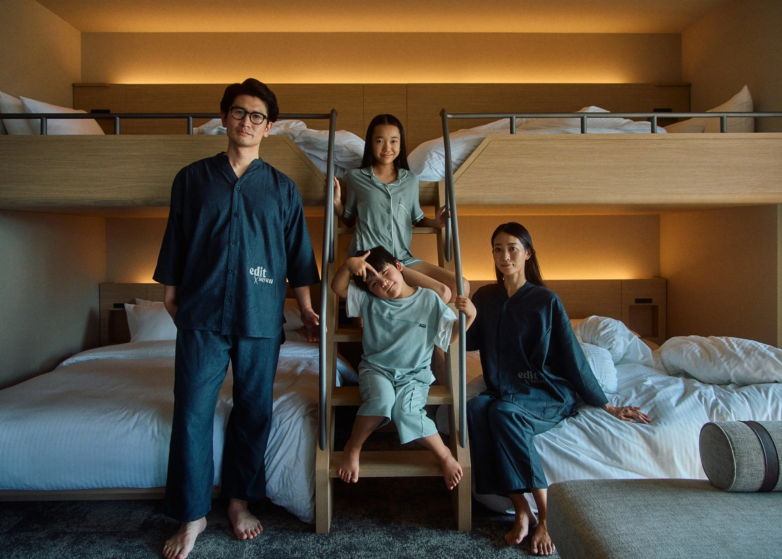 Family of four in pajamas standing and sitting on a staircase in a hotel room with two beds and warm lighting.
