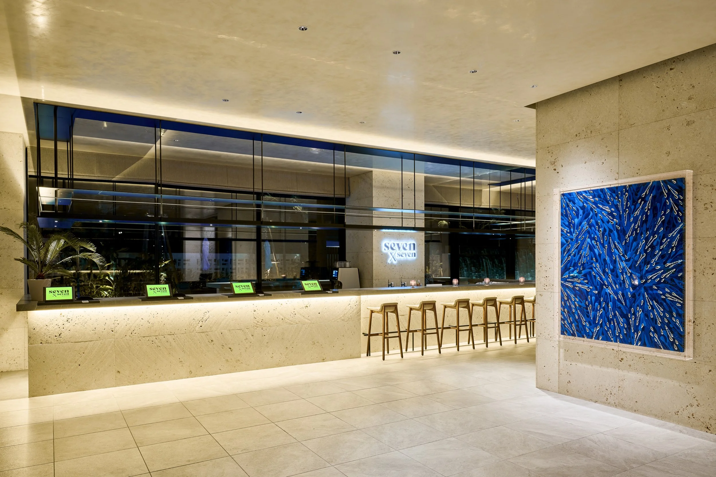 Modern hotel reception area with a long counter, five chairs, illuminated signage reading 'seven X seven', and abstract blue artwork on the wall.
