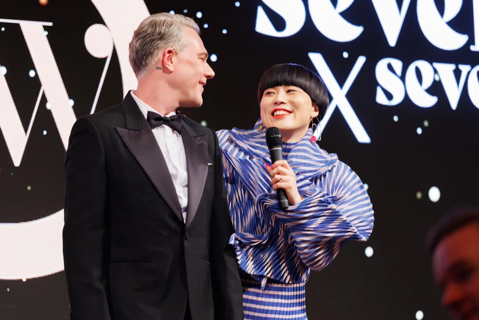 A man in a black tuxedo and a woman in a blue striped outfit are smiling at each other, standing on stage with a microphone. The woman holds the microphone and wears large hoop earrings. The background has a black screen with white illuminated text a