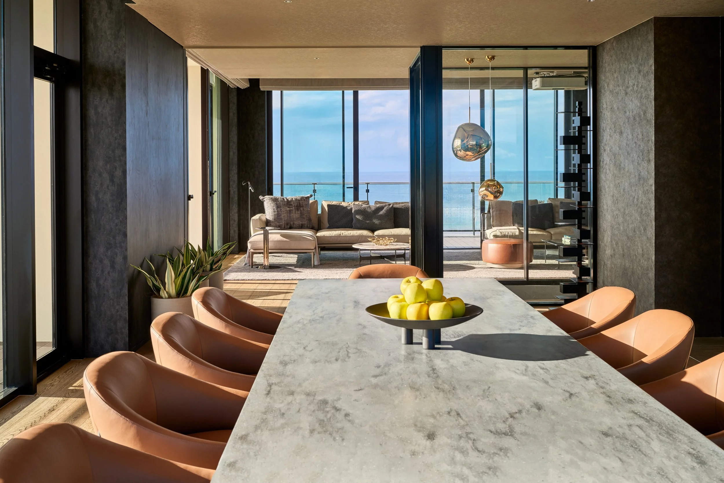 Modern dining area with a marble table surrounded by tan leather chairs. A bowl of apples is on the table. The room features large windows with a view of the ocean, a seating area with a sofa and chairs on a balcony, and contemporary lighting fixture