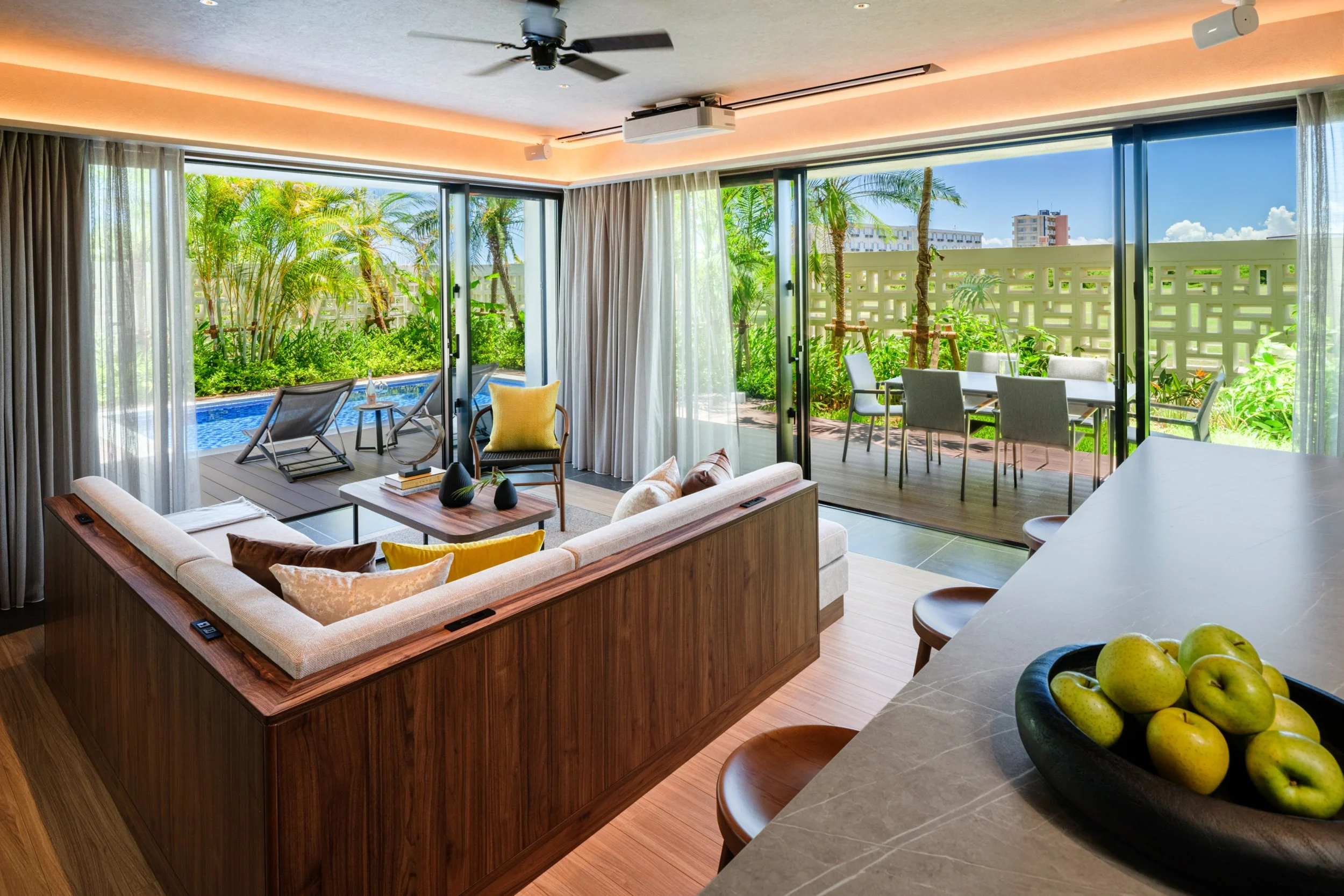 Living room with large sliding glass doors opening to a patio with outdoor furniture and greenery, kitchen counter with a bowl of green apples.