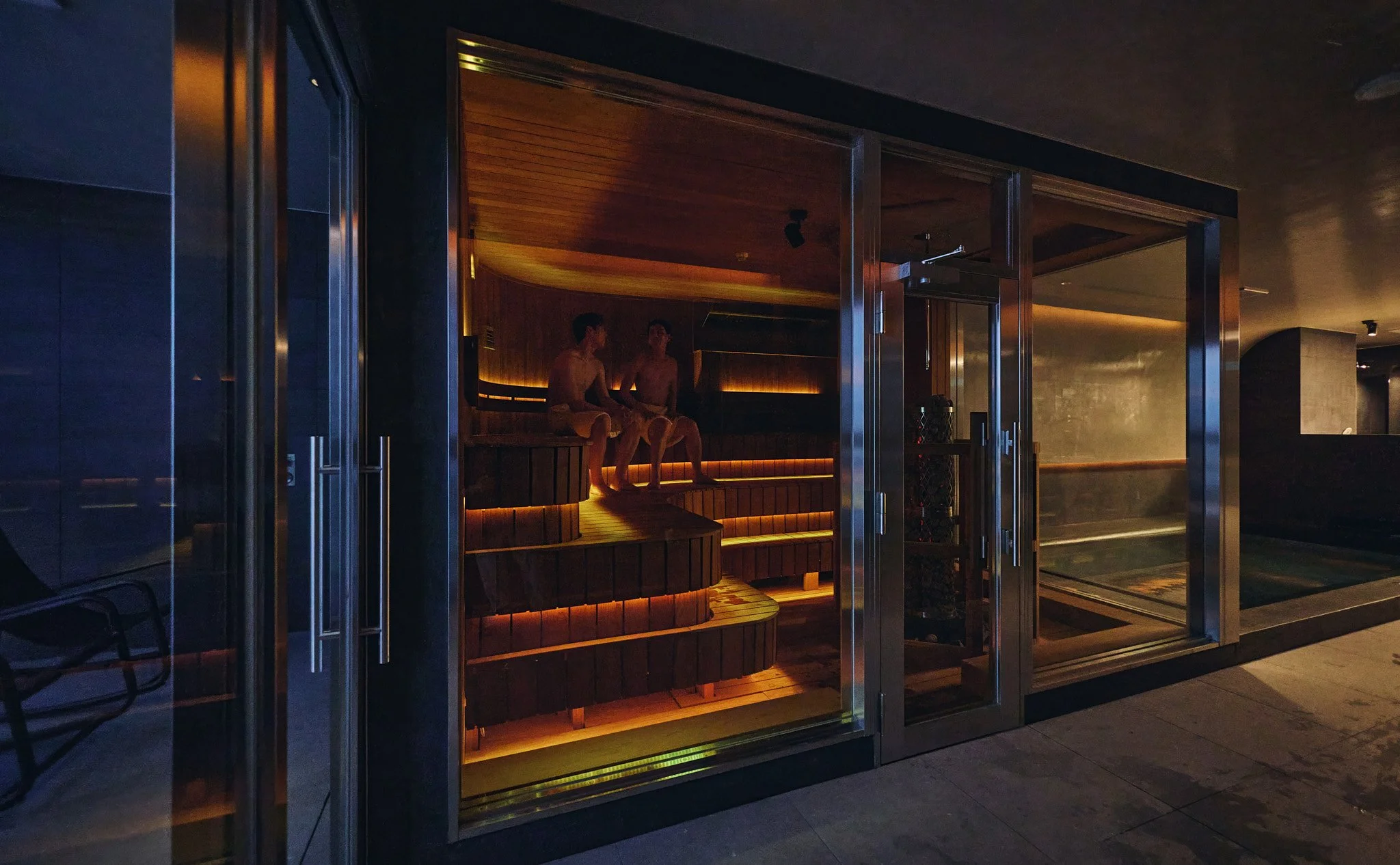 Two people sitting in a wooden sauna with warm lighting, visible through a glass door, next to a spa or pool area.