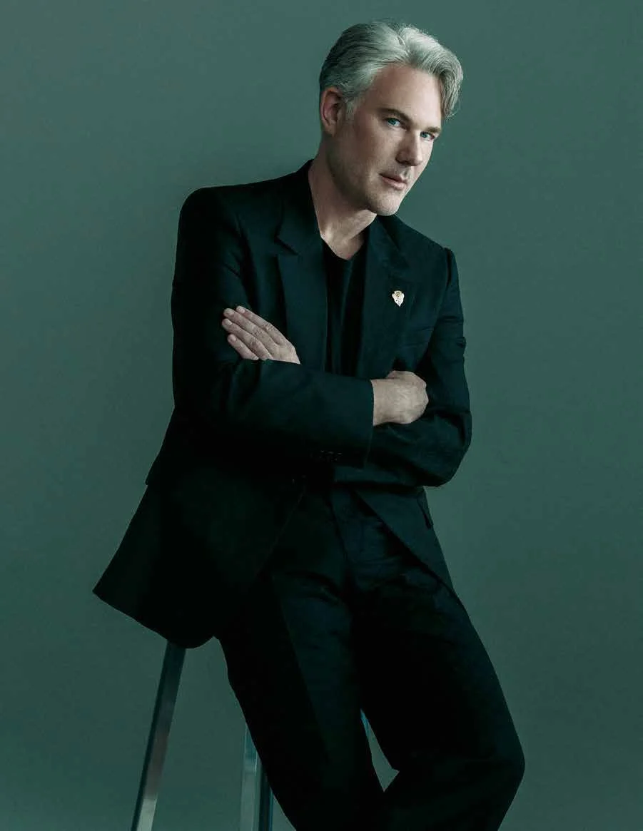 David Miskin in a black suit on a stool against a plain green background, crossing his arms and gazing at the camera.