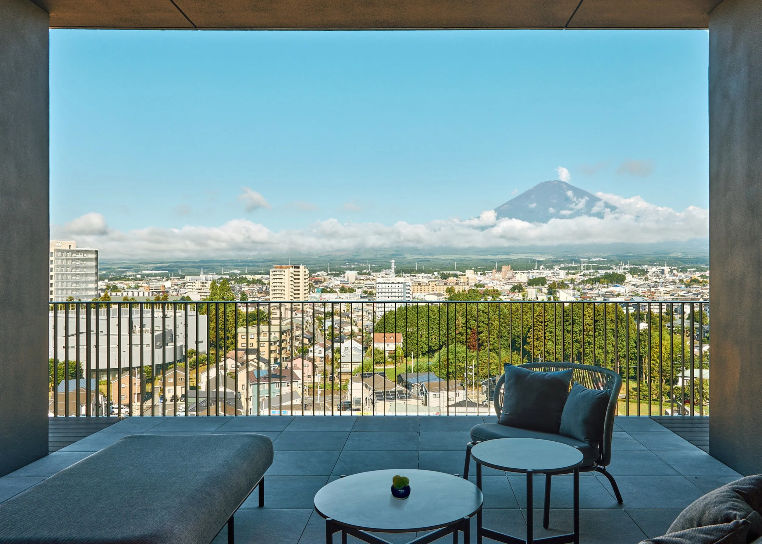 A balcony with a black railing overlooking a cityscape with tall buildings and trees. A large mountain with a cloud near its peak is visible in the distance. The balcony has outdoor furniture, including a cushioned sofa, a small round table with a sm