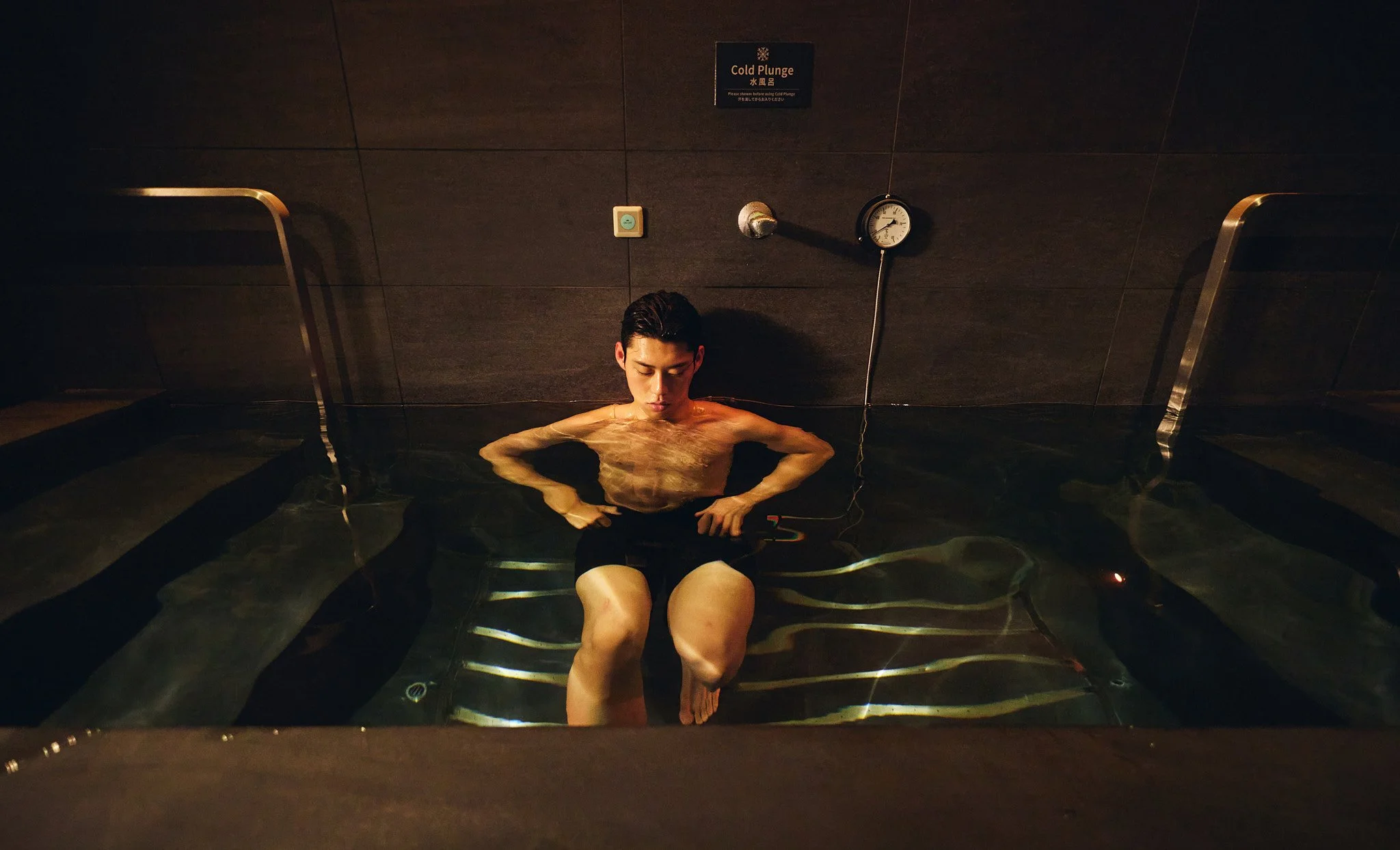 A man sitting in a dark tiled water pool with water reaching up to his chest, holding an electronic device, with a sign above that says 'Cold Plunge' and a water temperature gauge on the wall.