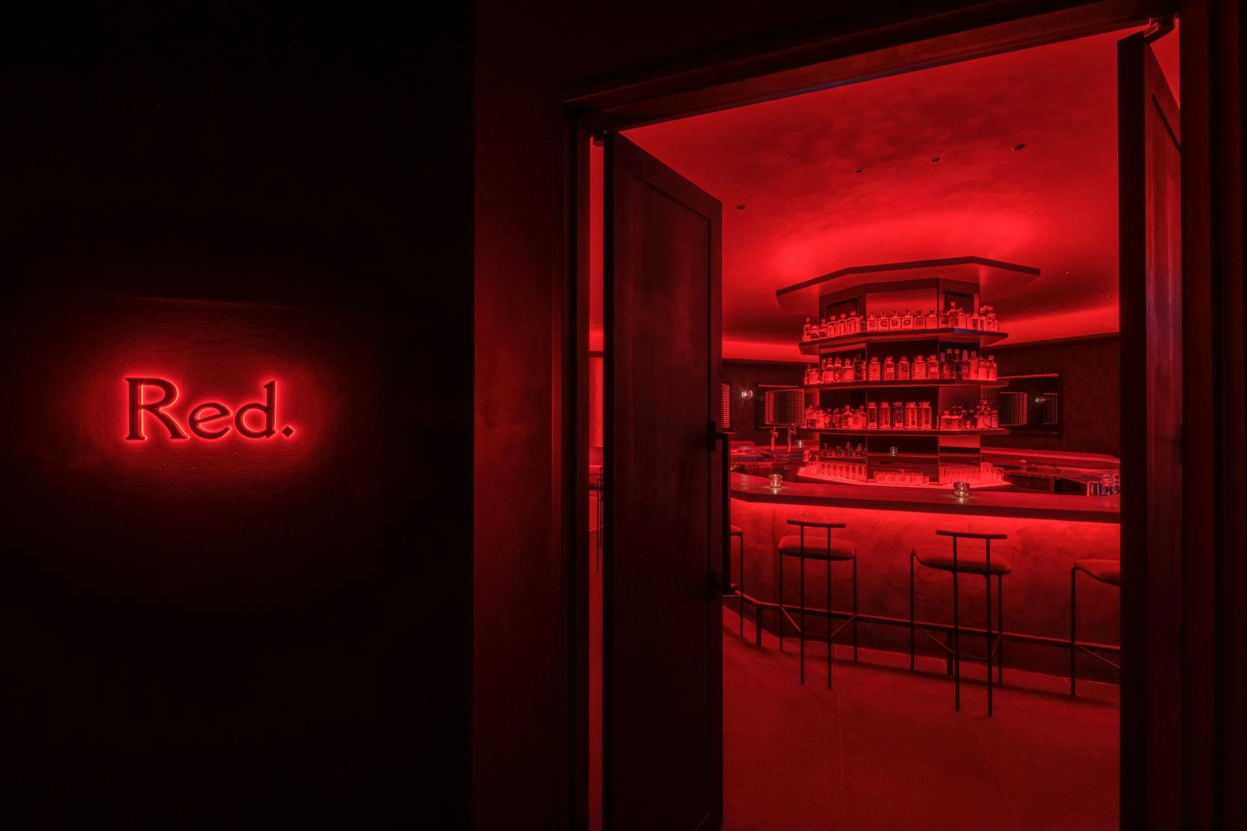 A dimly lit bar area with red lighting. The word 'Red.' is illuminated on a black wall.