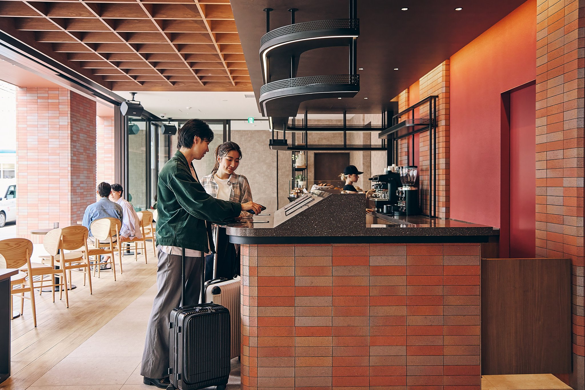 Two people with luggage ordering at a cafe counter inside a modern coffee shop with brick walls and seating area.