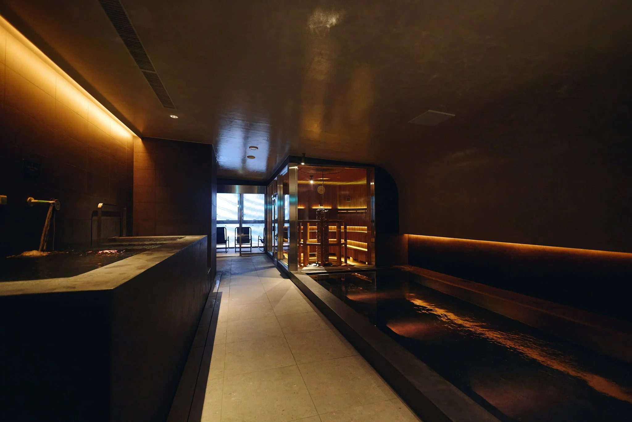 A modern, dimly lit spa room with a small water feature, wooden sauna, and seating area near a window with a view.